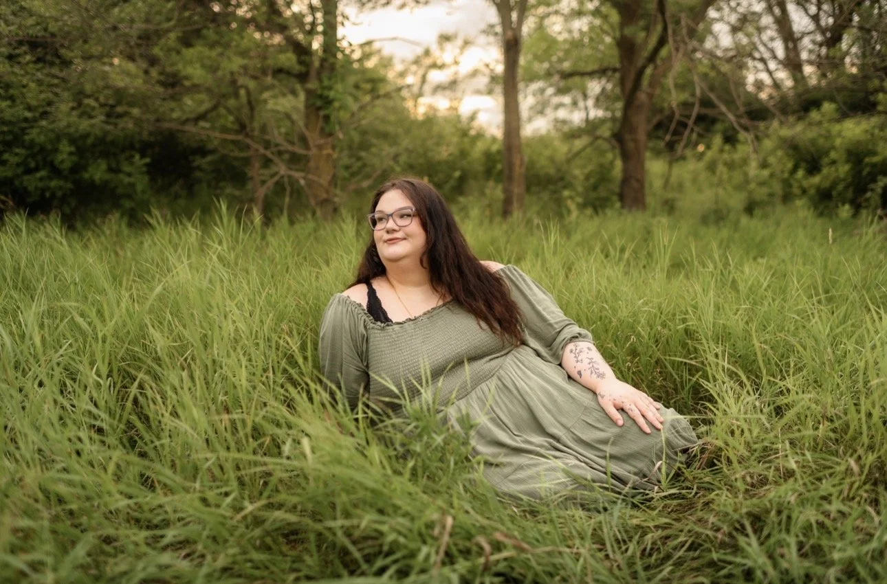 Woman lying in tall green grass outdoors during sunset, wearing a green dress and glasses, with tattoos on her arm.
