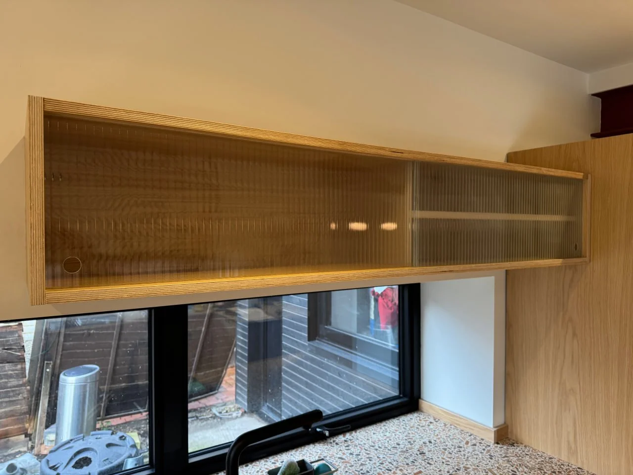 Wooden wall cabinet with glass doors mounted above a sliding glass door, with outdoor view of a fenced backyard and garden tools.