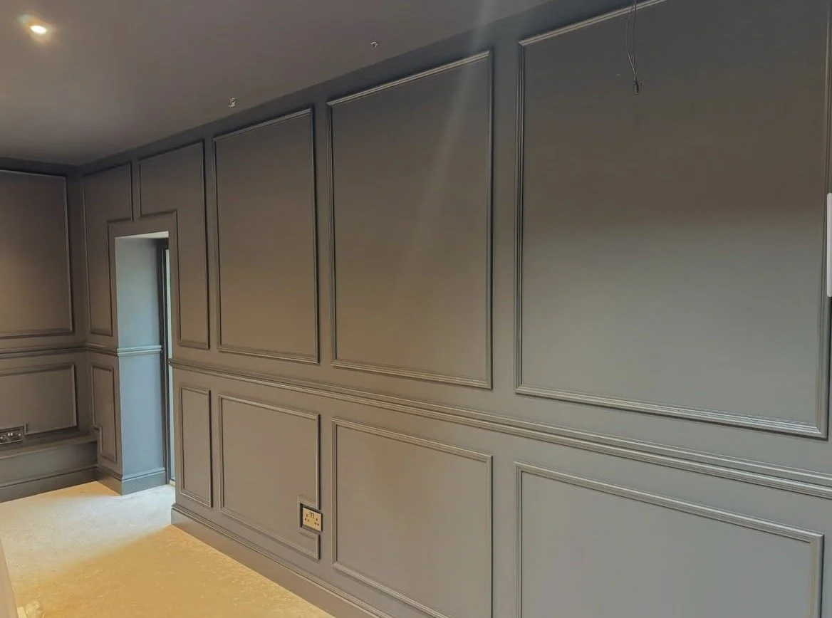 Interior of a room with gray paneled walls and a small door on the left. There is an outlet on the lower part of the wall and a hole in the ceiling with a hanging wire.