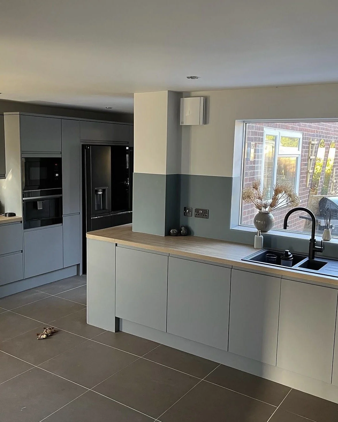 Modern kitchen with gray cabinets, black refrigerator, wooden countertop, window with a vase of dried flowers, and gray tiled floor.