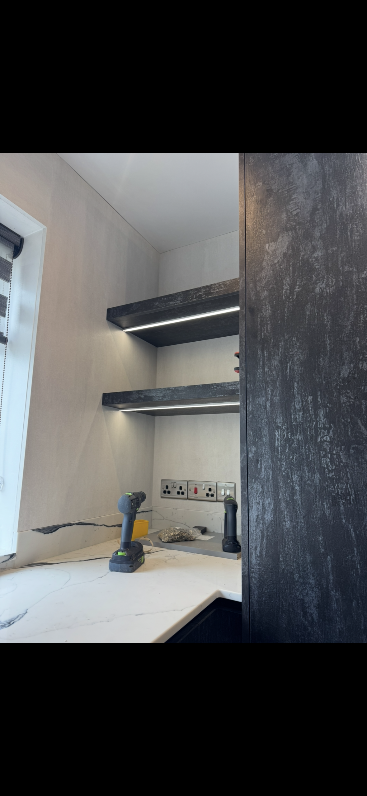 Under construction kitchen with dark floating shelves, electrical outlets, and cordless drills on a marble countertop.