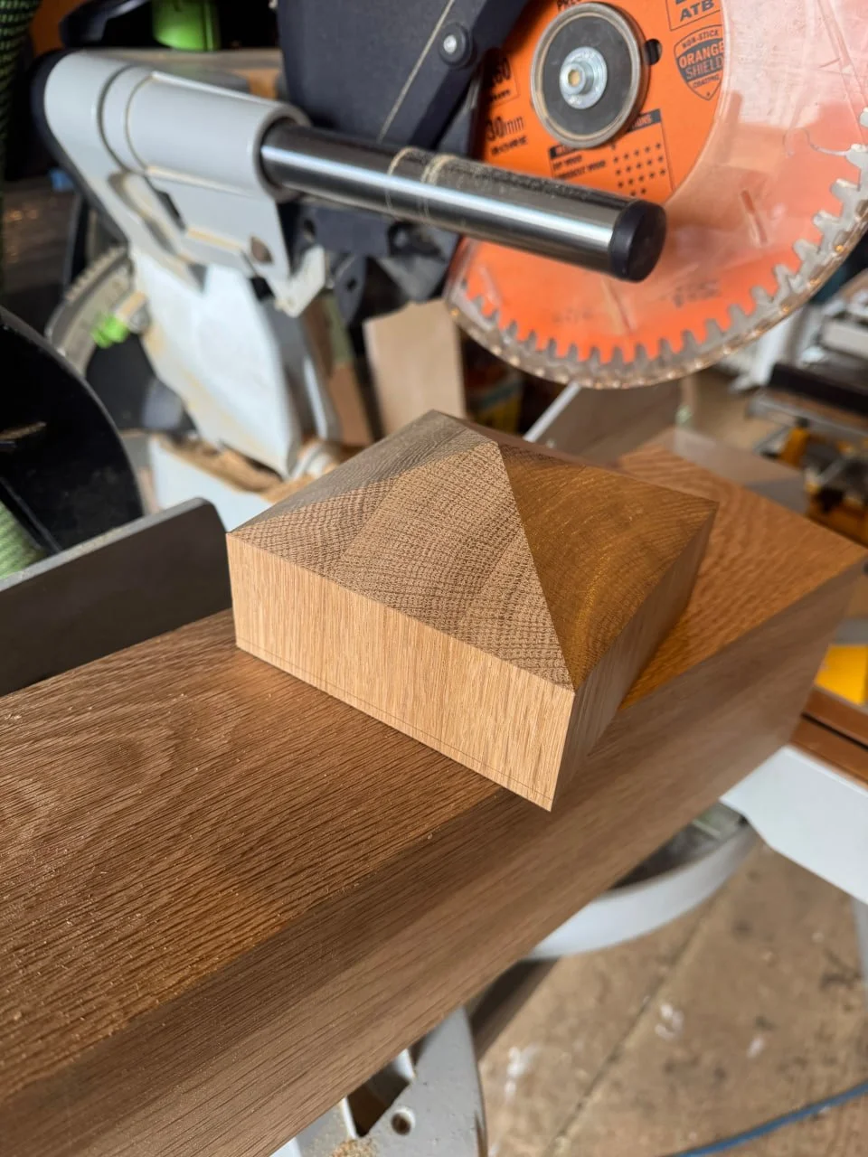 A wooden block with a carved pyramid shape on top is placed on a workbench, with a miter saw in the background.