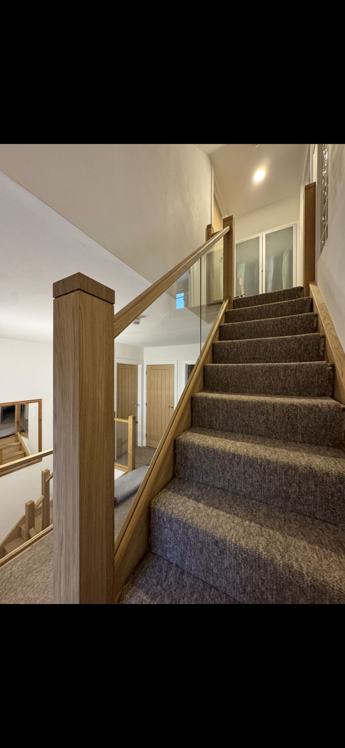 Interior view of a carpeted staircase with wooden handrails and glass panels, leading to an upper floor with closet doors and a hallway.