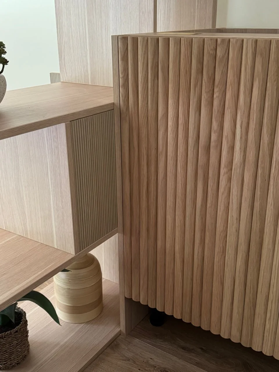 Close-up of wooden furniture with vertical slats, including a cabinet and shelves, in a room with wooden flooring.