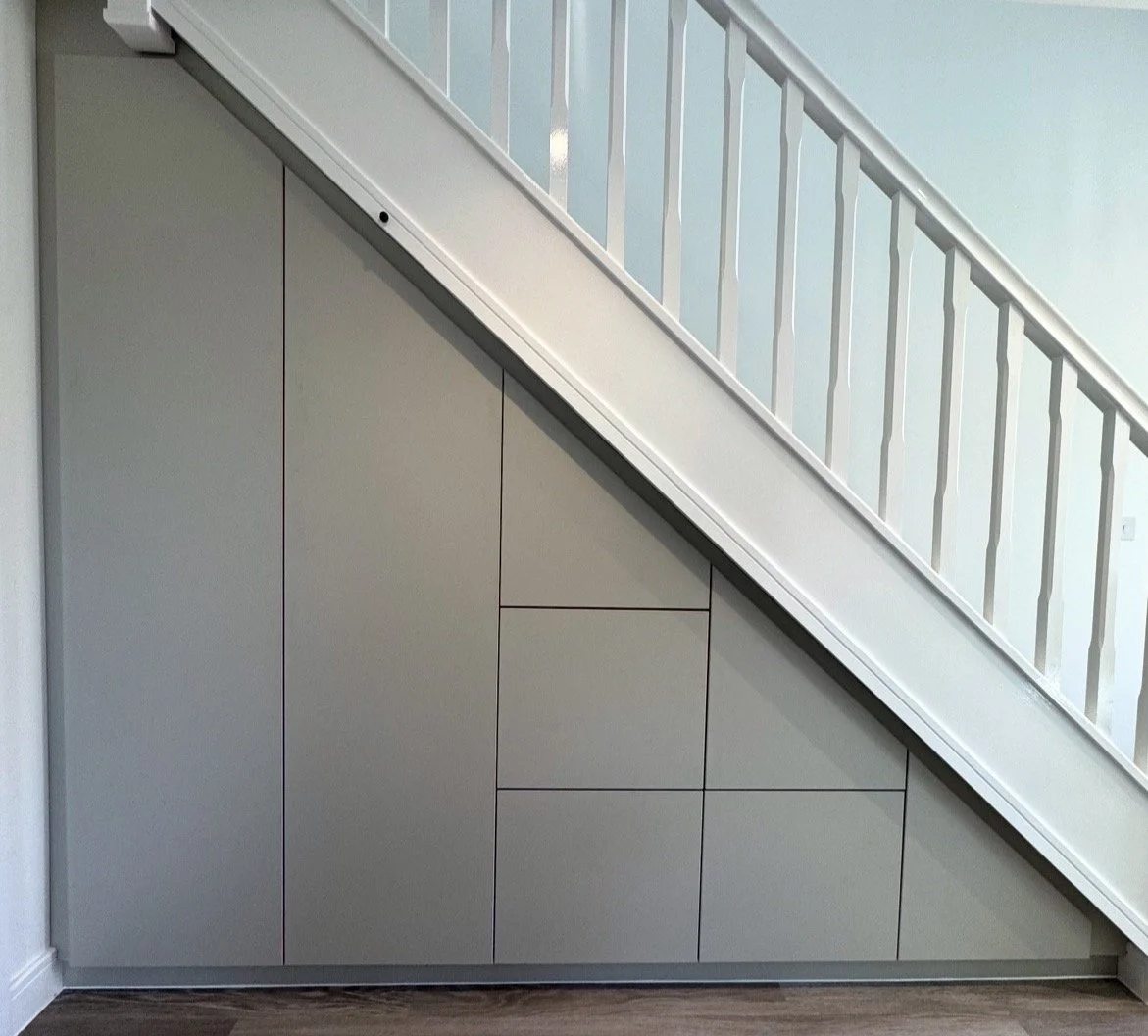 A built-in storage unit beneath a staircase with a modern, minimalist design, featuring gray panels and a white railing.