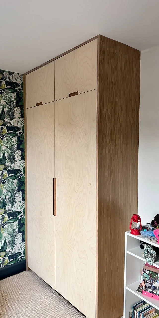 A tall wooden wardrobe with light-colored doors in a room with tropical-patterned wallpaper on one wall and a white shelf with children's toys on the side.