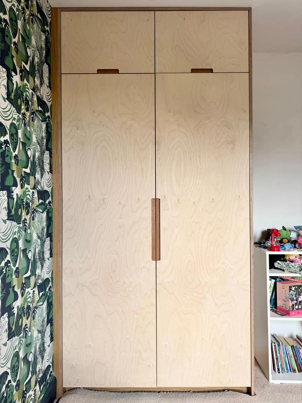 A tall, unfinished wooden closet with four doors and a vertical handle in the center. To the left is a patterned wallpaper wall with green, black, and white abstract designs. To the right, a white shelf with toys and books is partially visible.