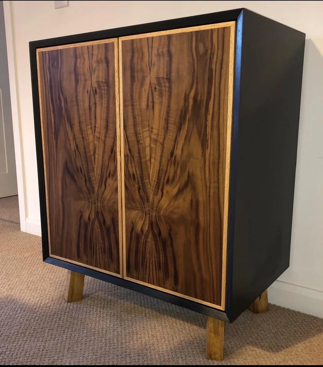 A wooden cabinet with sliding doors featuring distinctive wood grain patterns, supported by four angled wooden legs, placed on a carpeted floor indoors.