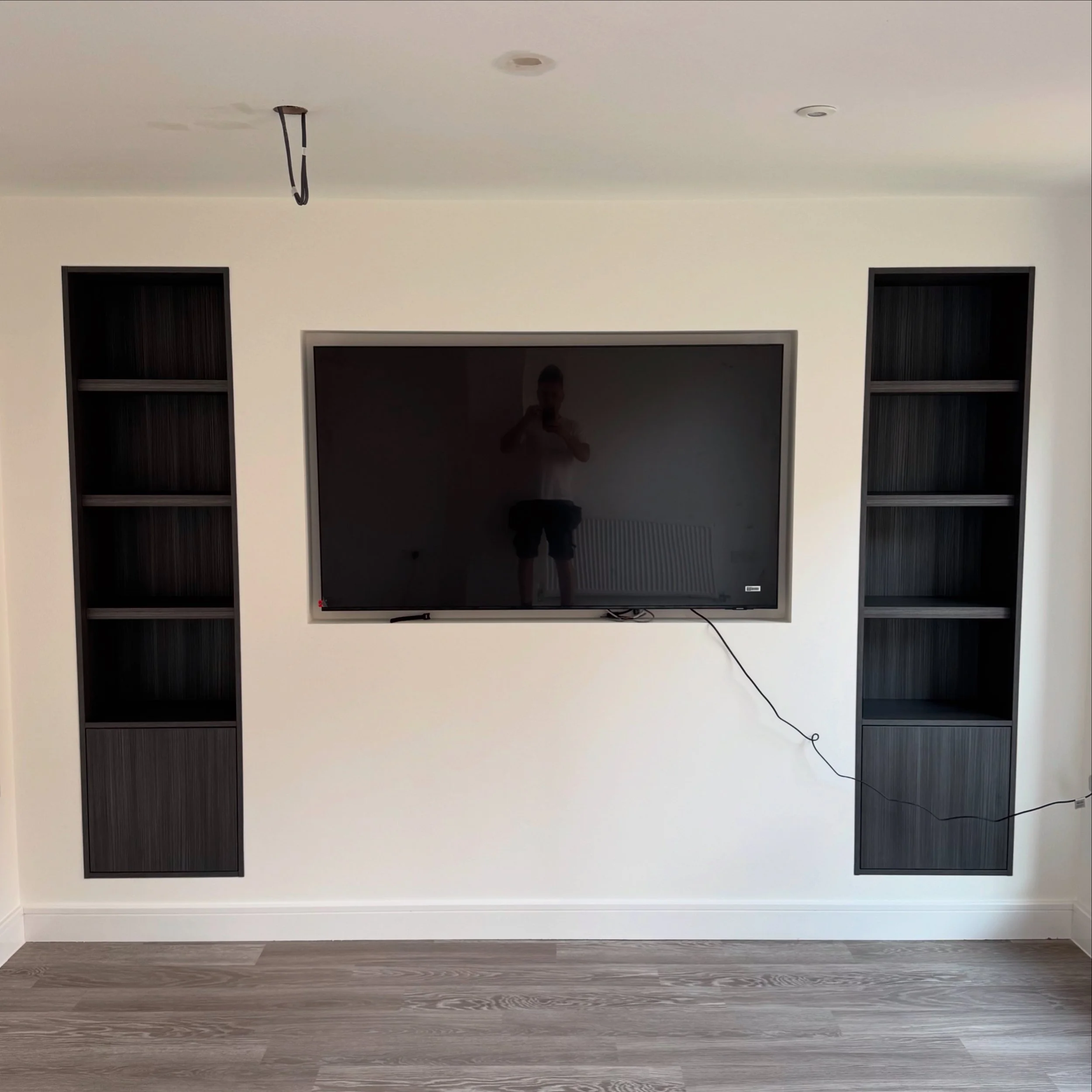 Interior wall with a flat screen TV and black shelving on either side, a person is visible in the TV's reflection.