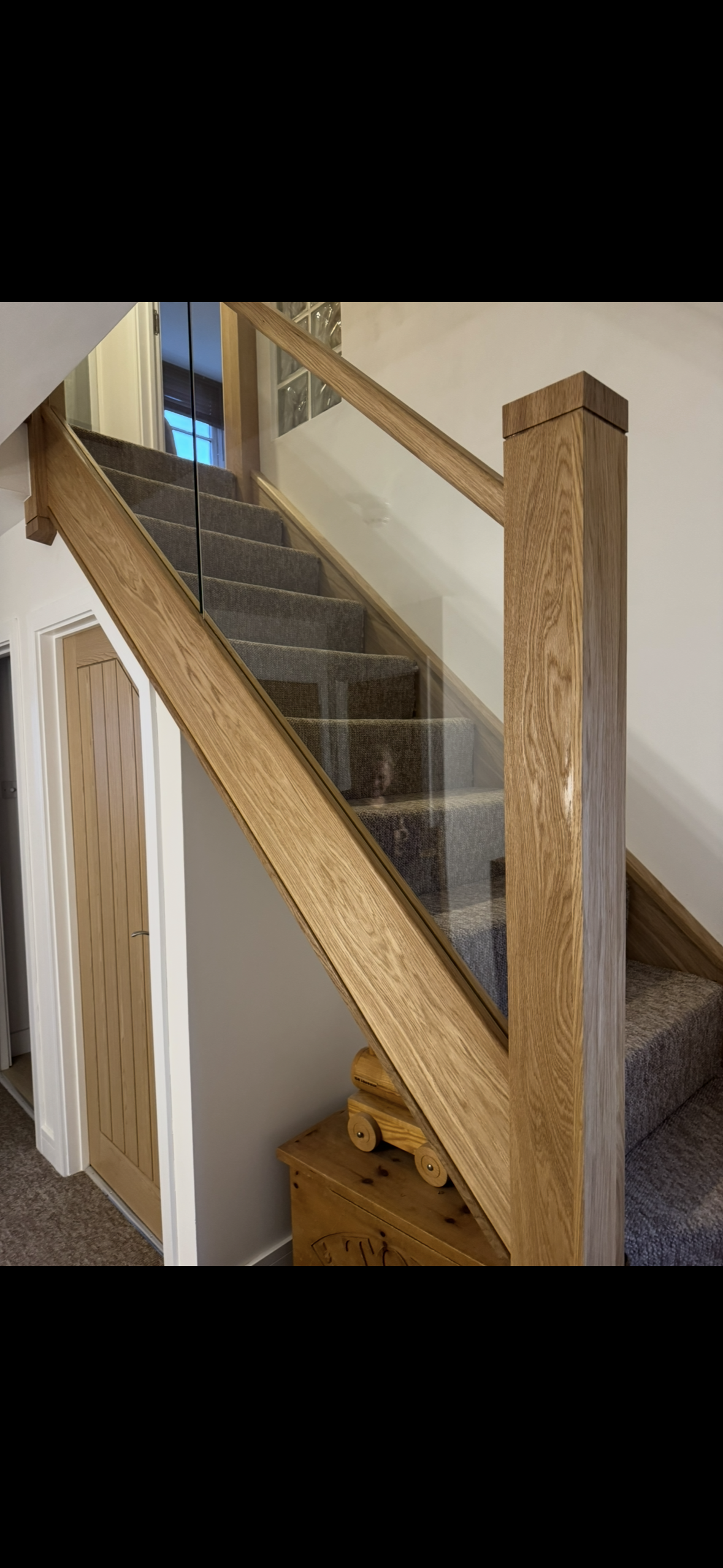 Interior staircase with wooden handrail and glass panels, carpeted stairs, and a corner with a closed wooden door and a small wooden storage chest.