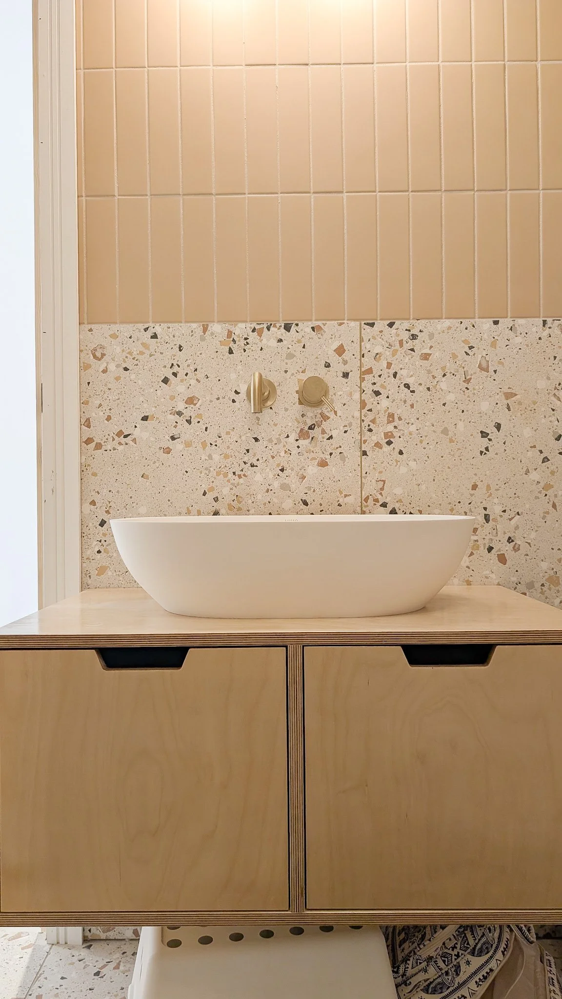 A modern bathroom vanity with a white vessel sink, beige terrazzo and tile wall behind, and a wooden cabinet with black handles below.
