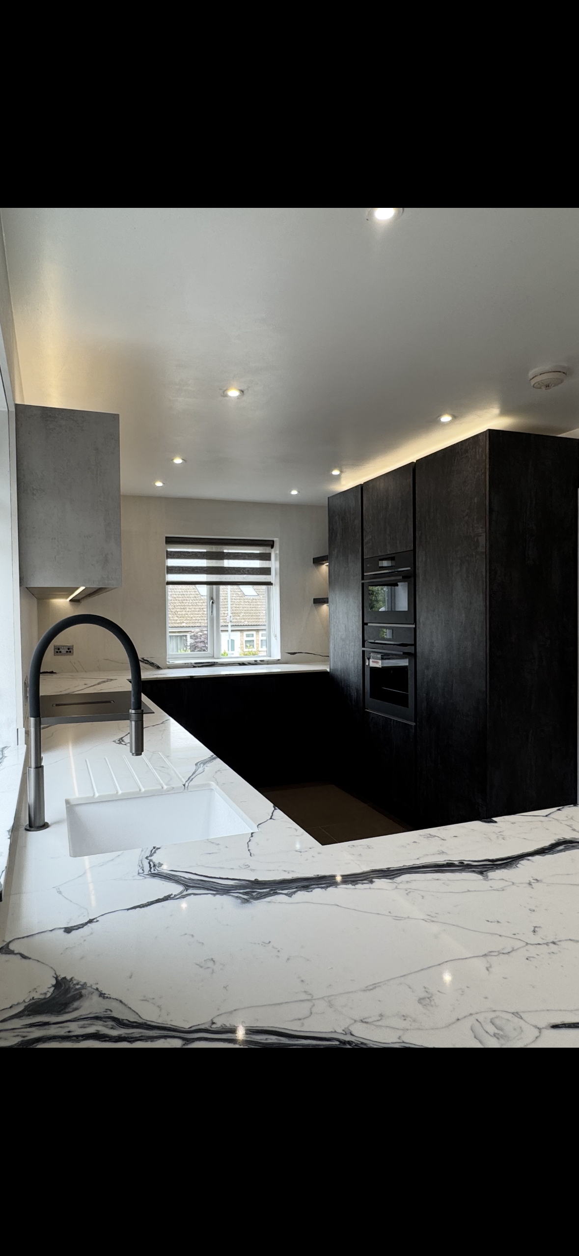 Modern kitchen with white marble countertop, black cabinets, and a window with blinds.