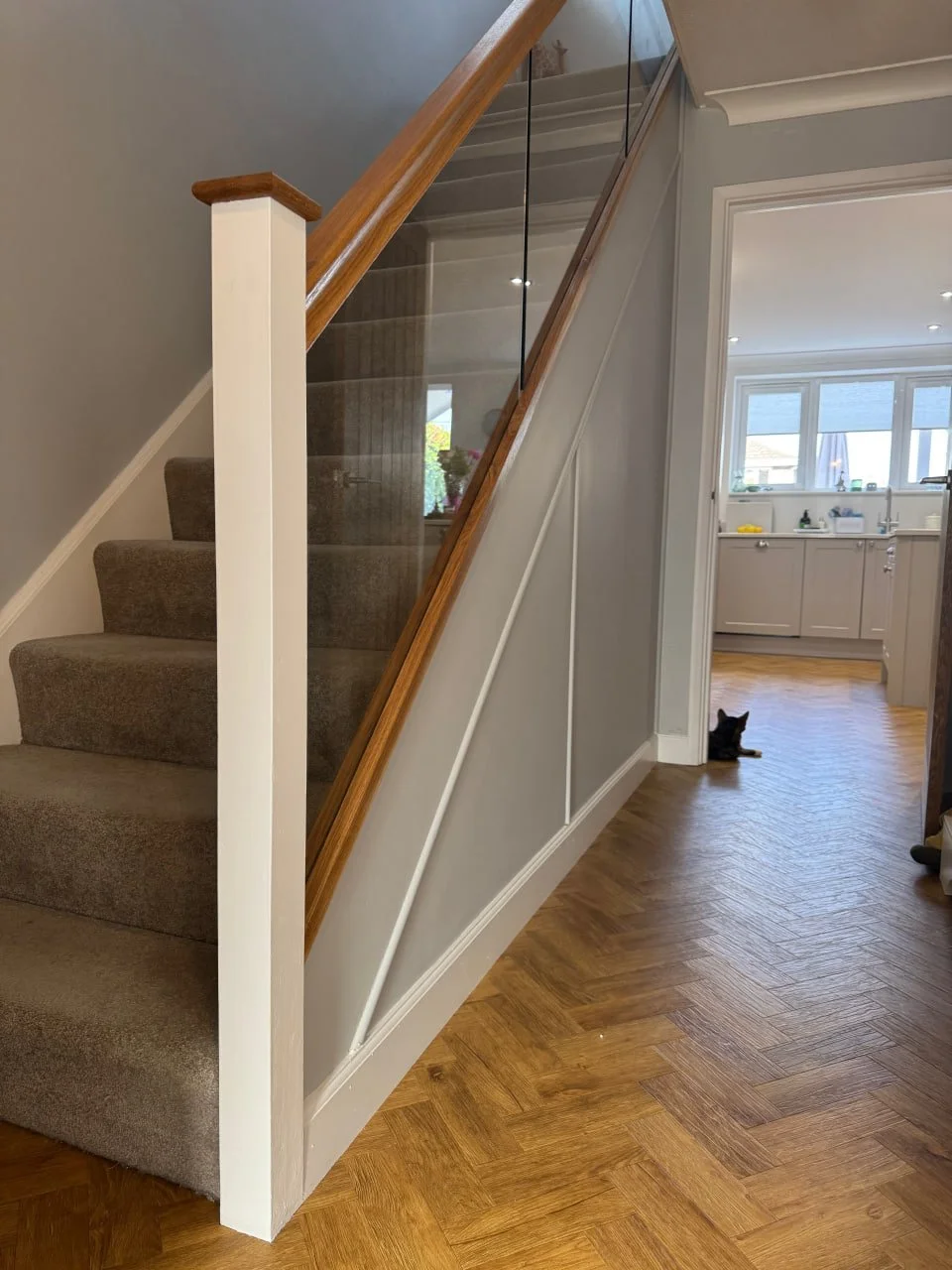 Interior view of a staircase with brown carpet, wooden handrail, and glass panel, beside a white wall with decorative molding, leading to a kitchen with wooden floor and a black cat sitting near the doorway.