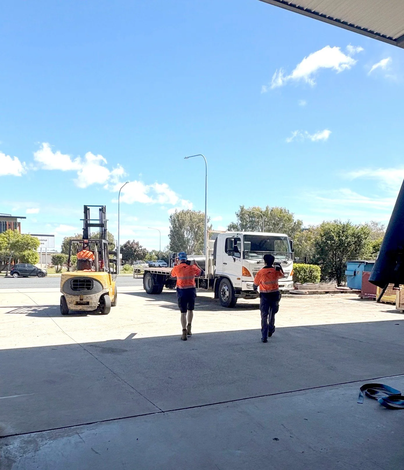 Three construction workers in orange safety shirts walk out from a building onto a parking lot, with a forklift, a white truck, and some trees in the background under a partly cloudy blue sky.