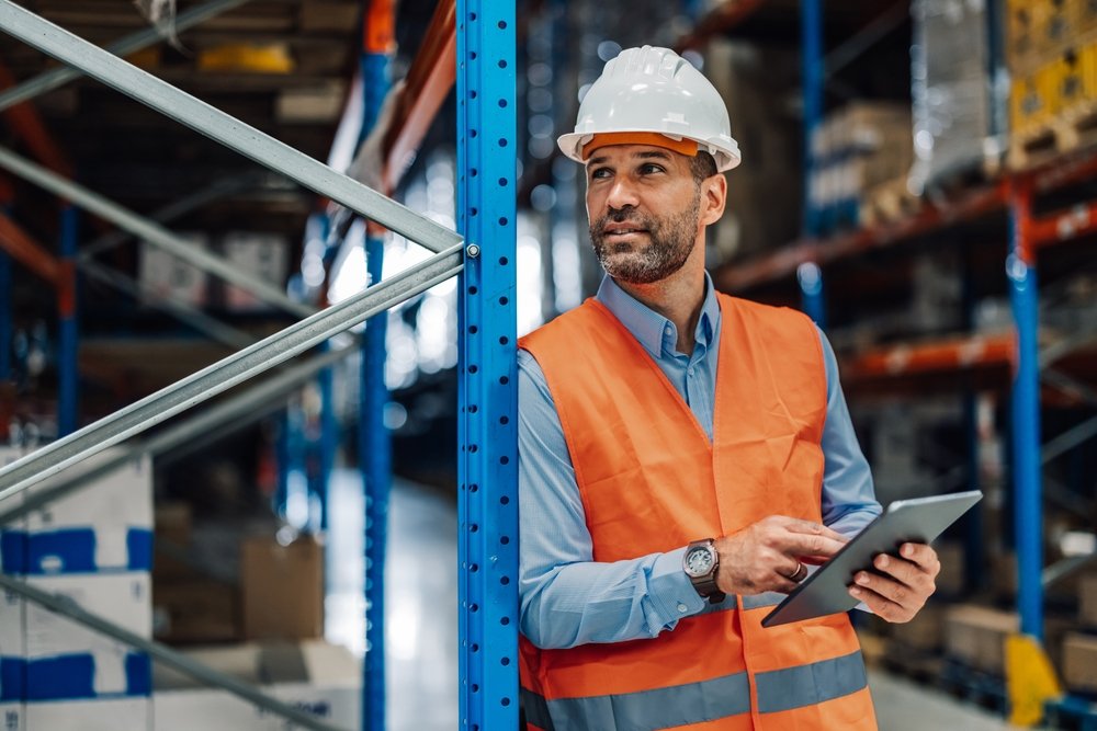 A man wearing a white hard hat and orange safety vest holding a tablet in a warehouse or storage facility.