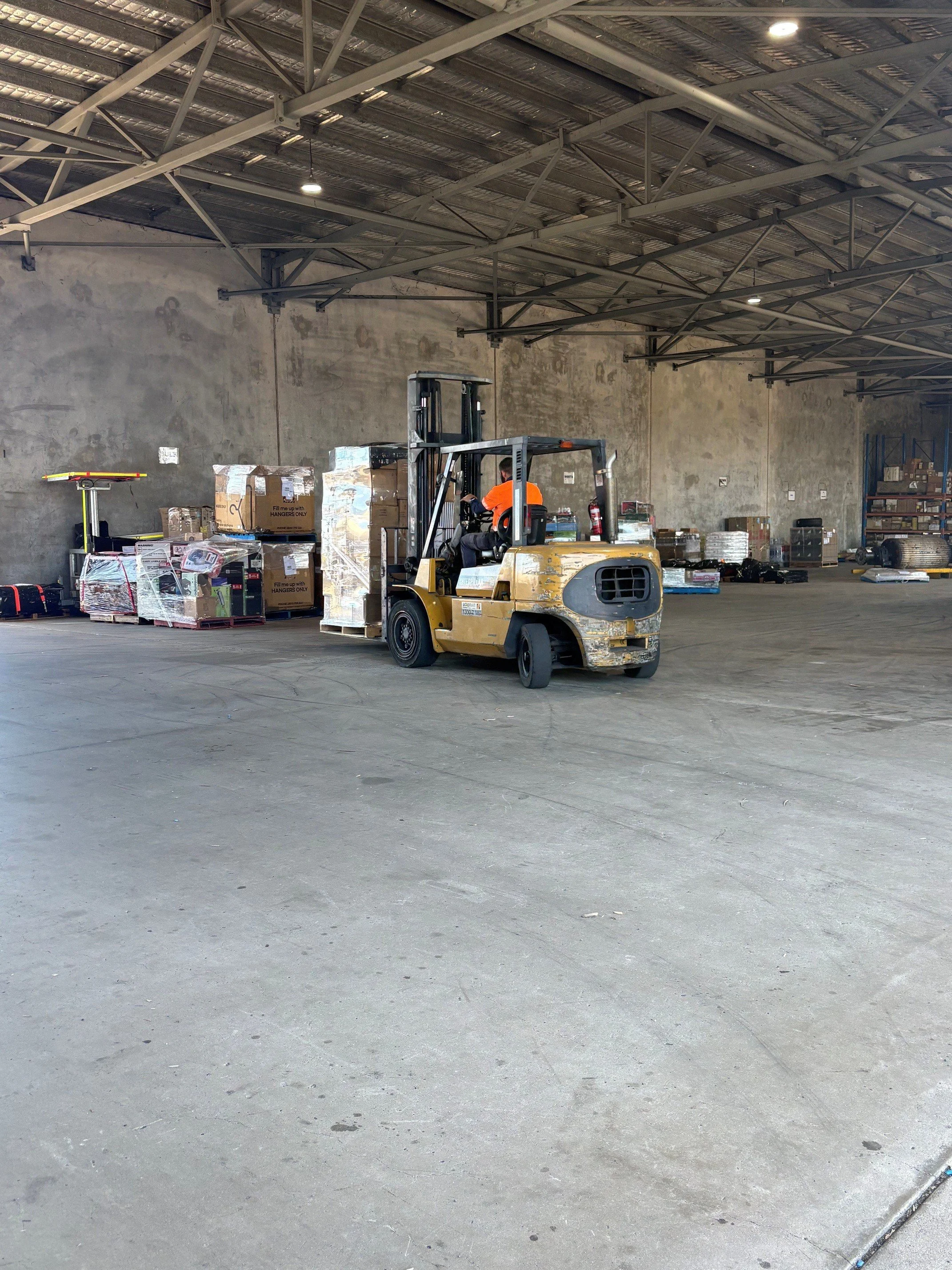Interior of a warehouse with a forklift moving pallets and boxes. The warehouse has high ceilings with metal beams and a concrete floor.