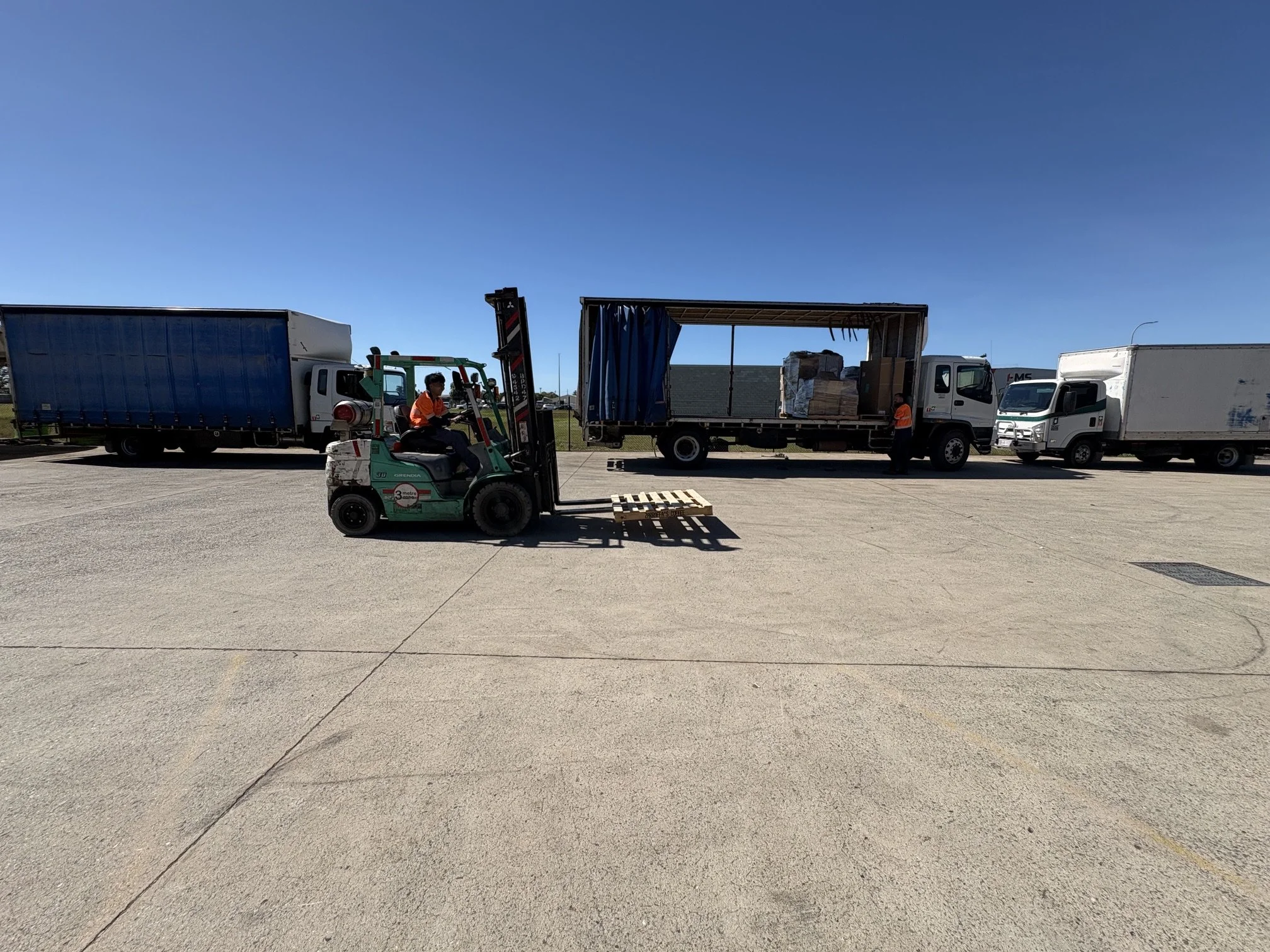 A forklift operator moves pallets of goods in front of two parked delivery trucks on a concrete lot under a clear blue sky.