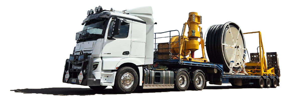 A white semi-truck hauling industrial equipment, including a large spool of cable or hose, against a plain background.