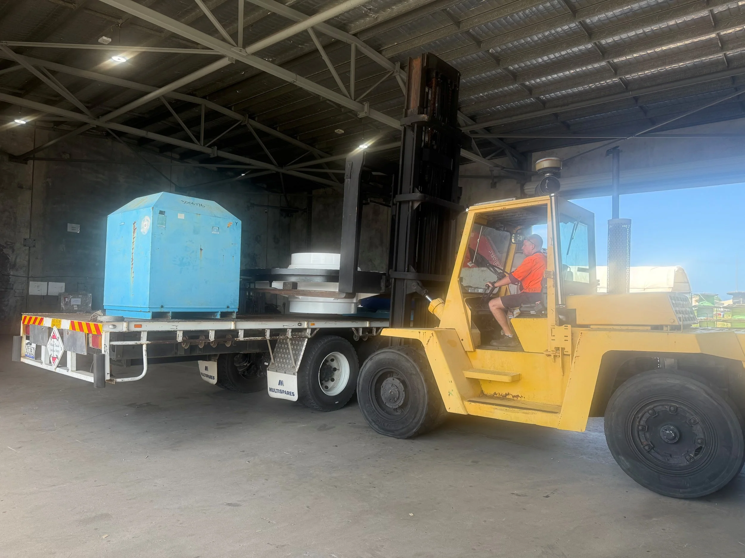 A worker operating a yellow forklift inside a warehouse, transporting large blue equipment on a flatbed truck.