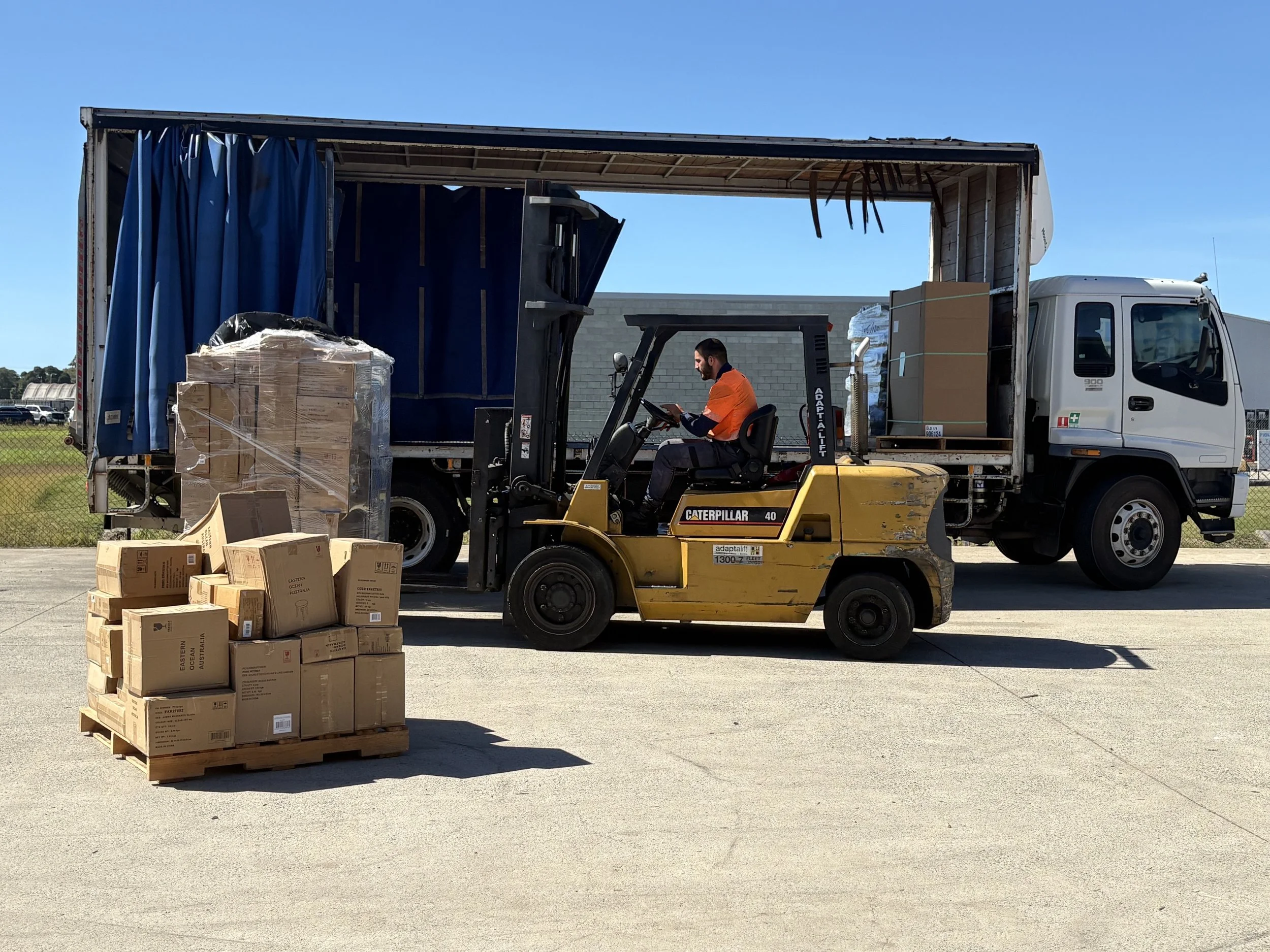 A man operating a forklift on a paved outdoor area, moving boxes from a large delivery truck with its side open, with a stack of boxes nearby, under a clear blue sky.
