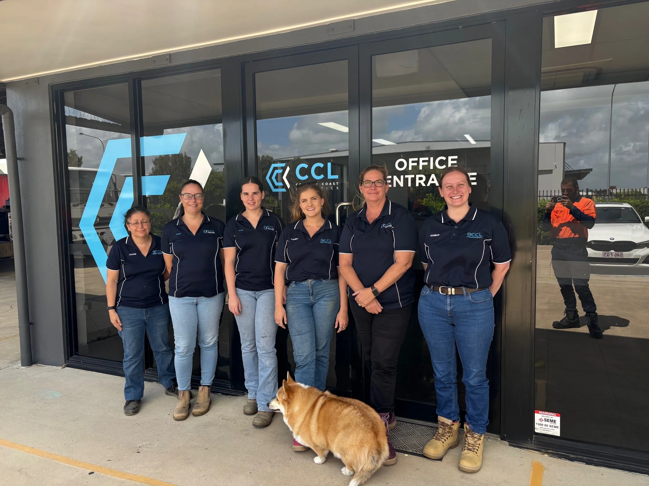 Group of six women standing outside in front of a glass door with the logo 'CCL Coast Logistics' and staff name tags, along with a brown and white dog in front of them. Reflection shows an outdoor area with cars and a person in an orange vest.