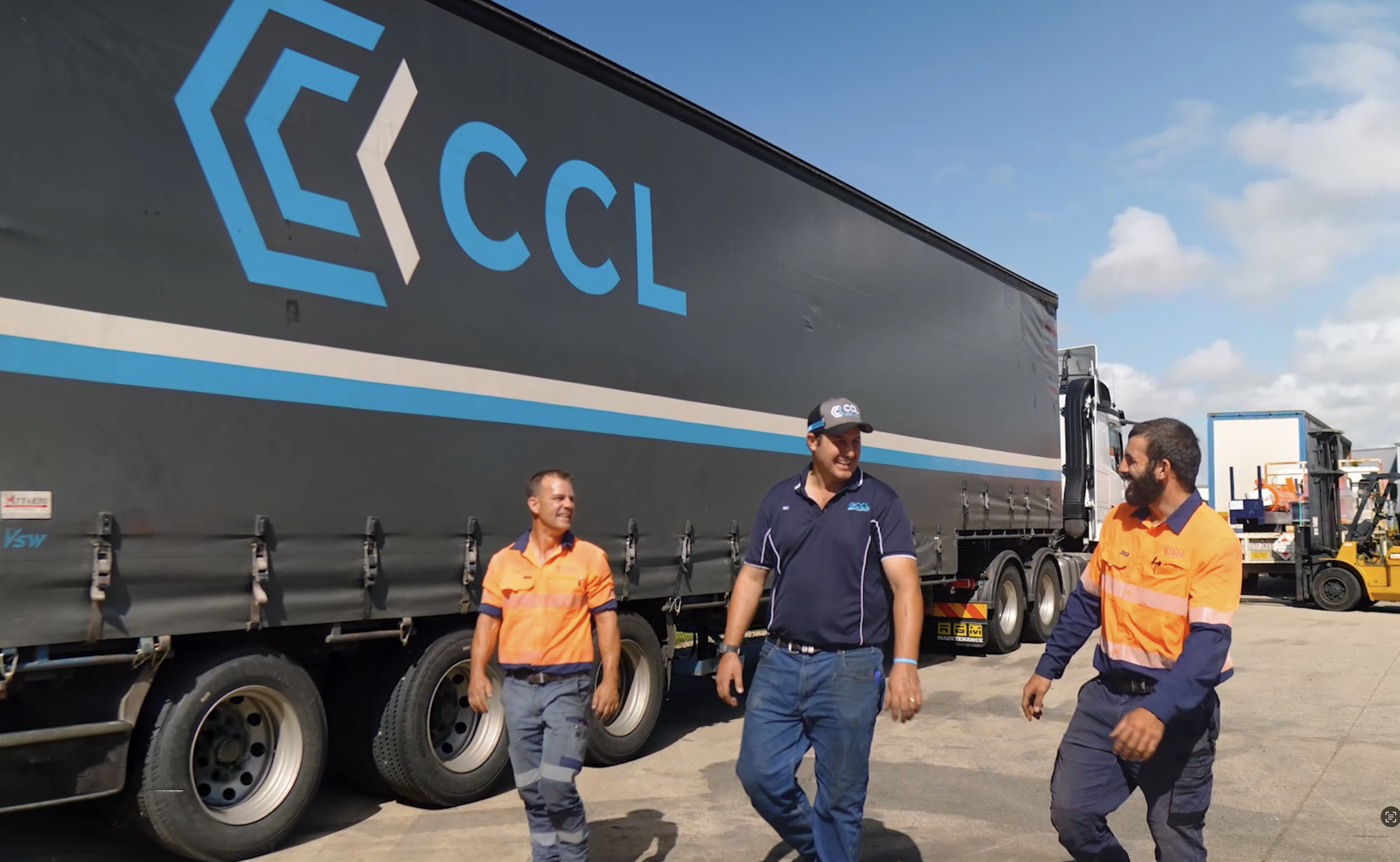 Four men are standing and walking near a large truck with the logo 'CCL' on its side. Two men are dressed in orange safety uniforms, and the other two in casual dark clothing. The background shows a bright blue sky with some clouds.