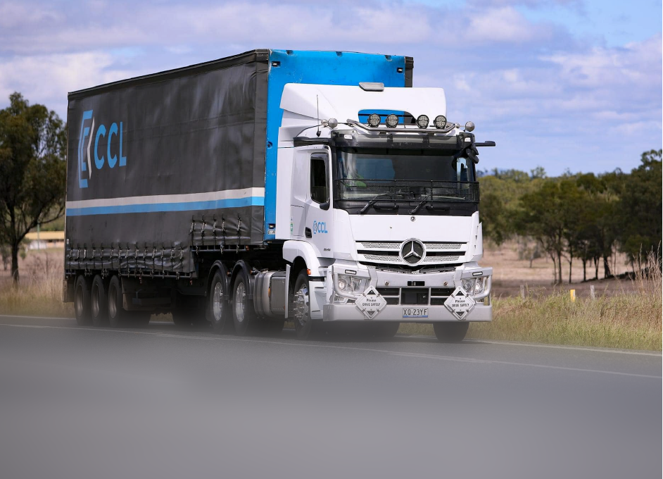 A transport truck with a black and blue CCL trailer driving on a rural road with trees in the background.