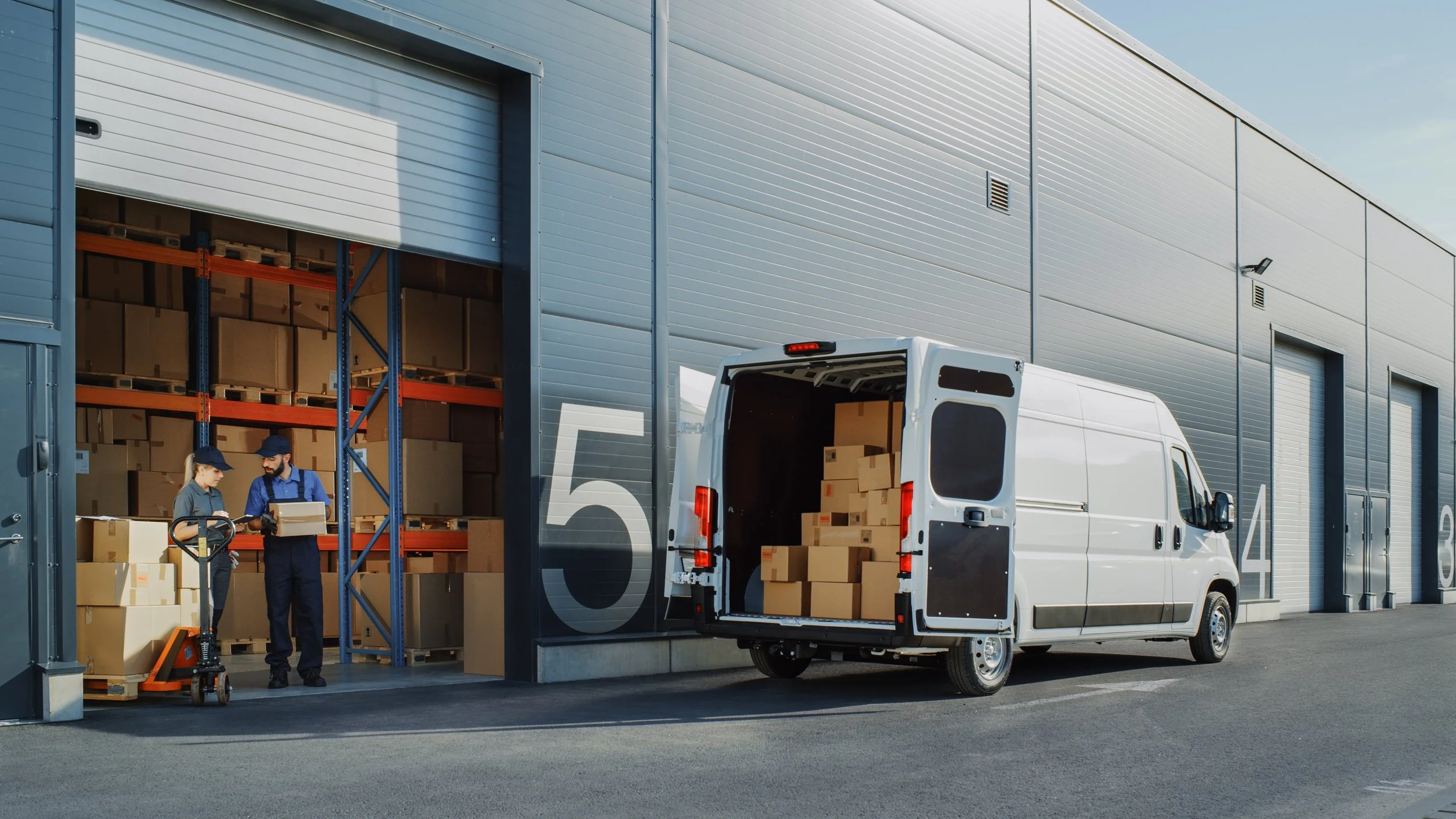 Workers loading boxes into a delivery van outside a large warehouse with a partially open garage door.