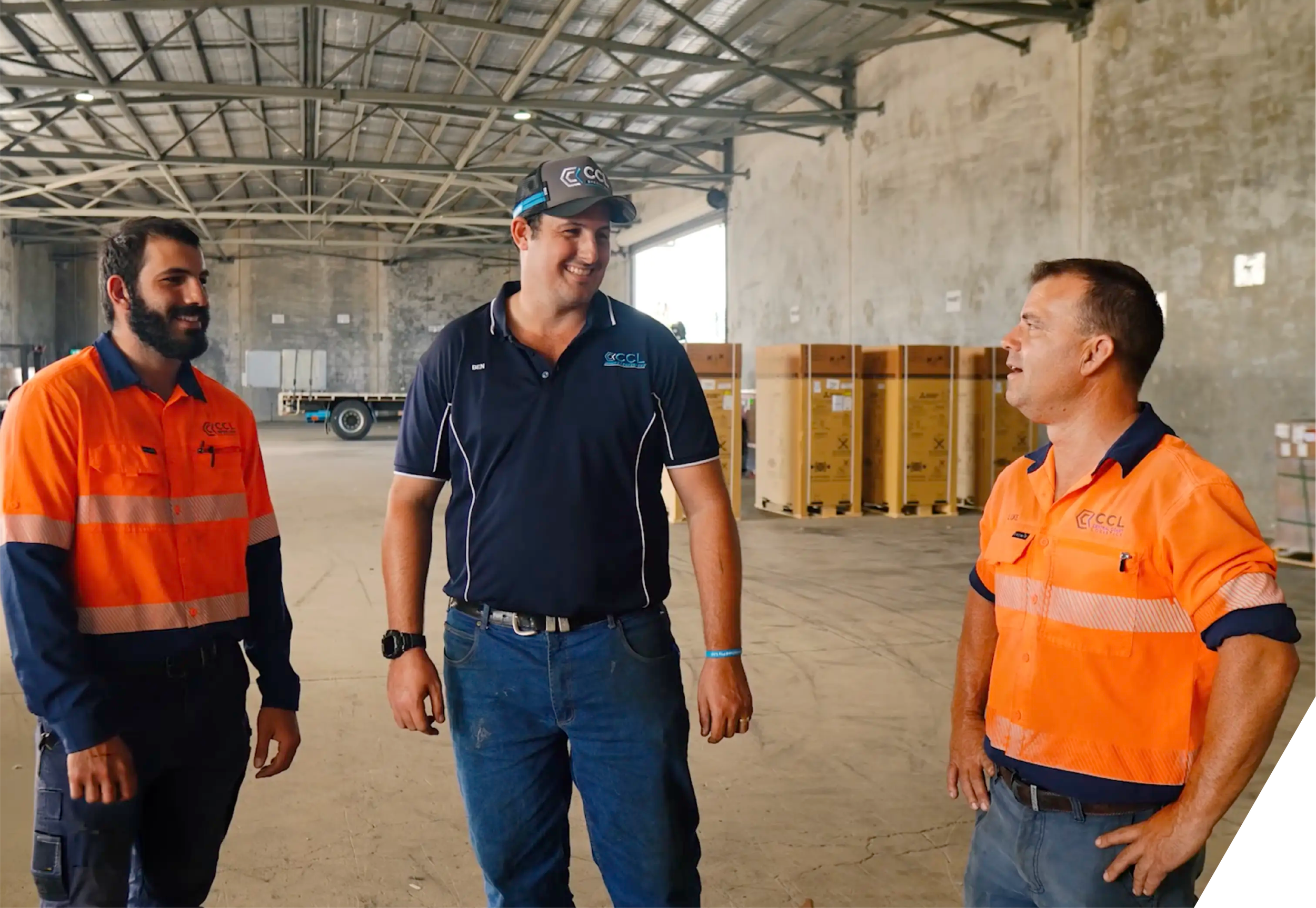 Three men in a spacious industrial warehouse are engaged in conversation. Two are wearing bright orange safety uniforms, and one is dressed in a navy blue polo shirt with a logo. They are smiling and standing near cardboard boxes on pallets. The background features metal beams and an open ceiling.