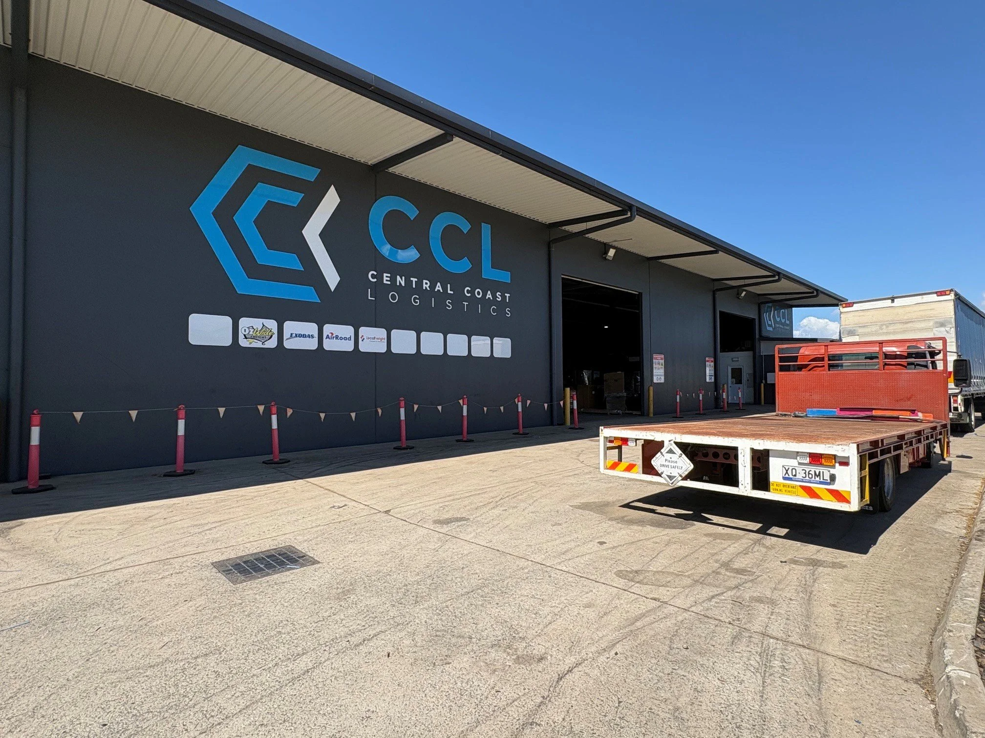 Exterior of a large warehouse with a logo for Central Coast Logistics, a list of partner companies, and a loading dock area with trucks and a flatbed trailer.