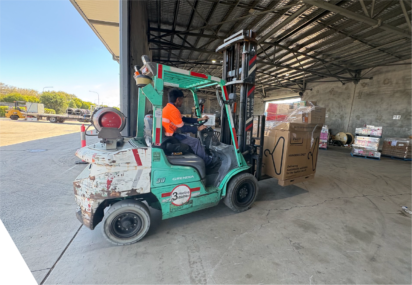 A worker in an orange safety vest operates a green forklift in a warehouse, lifting a large cardboard box .