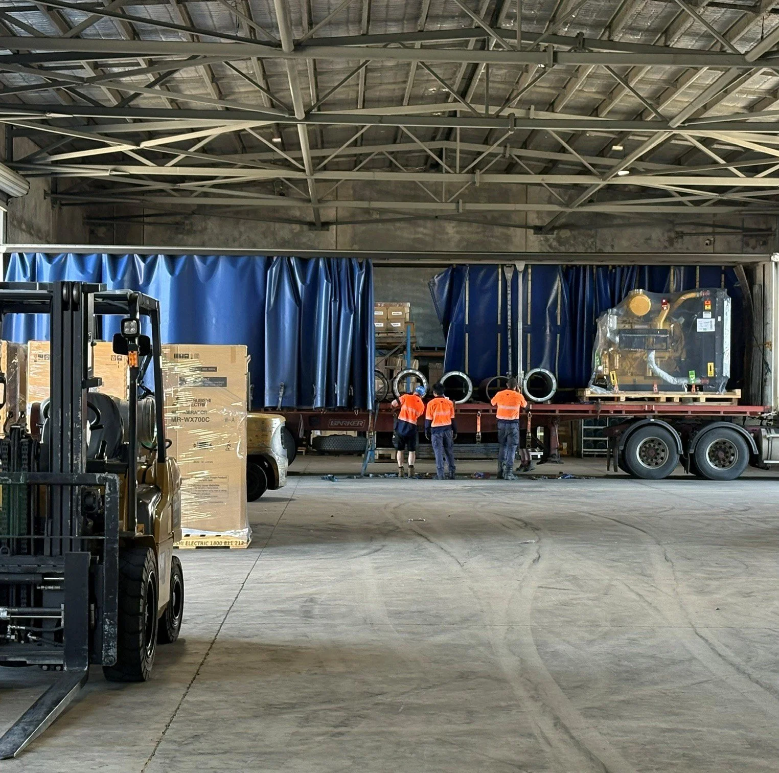 Four workers in orange shirts loading or unloading equipment and cargo onto a large flatbed truck inside a warehouse.