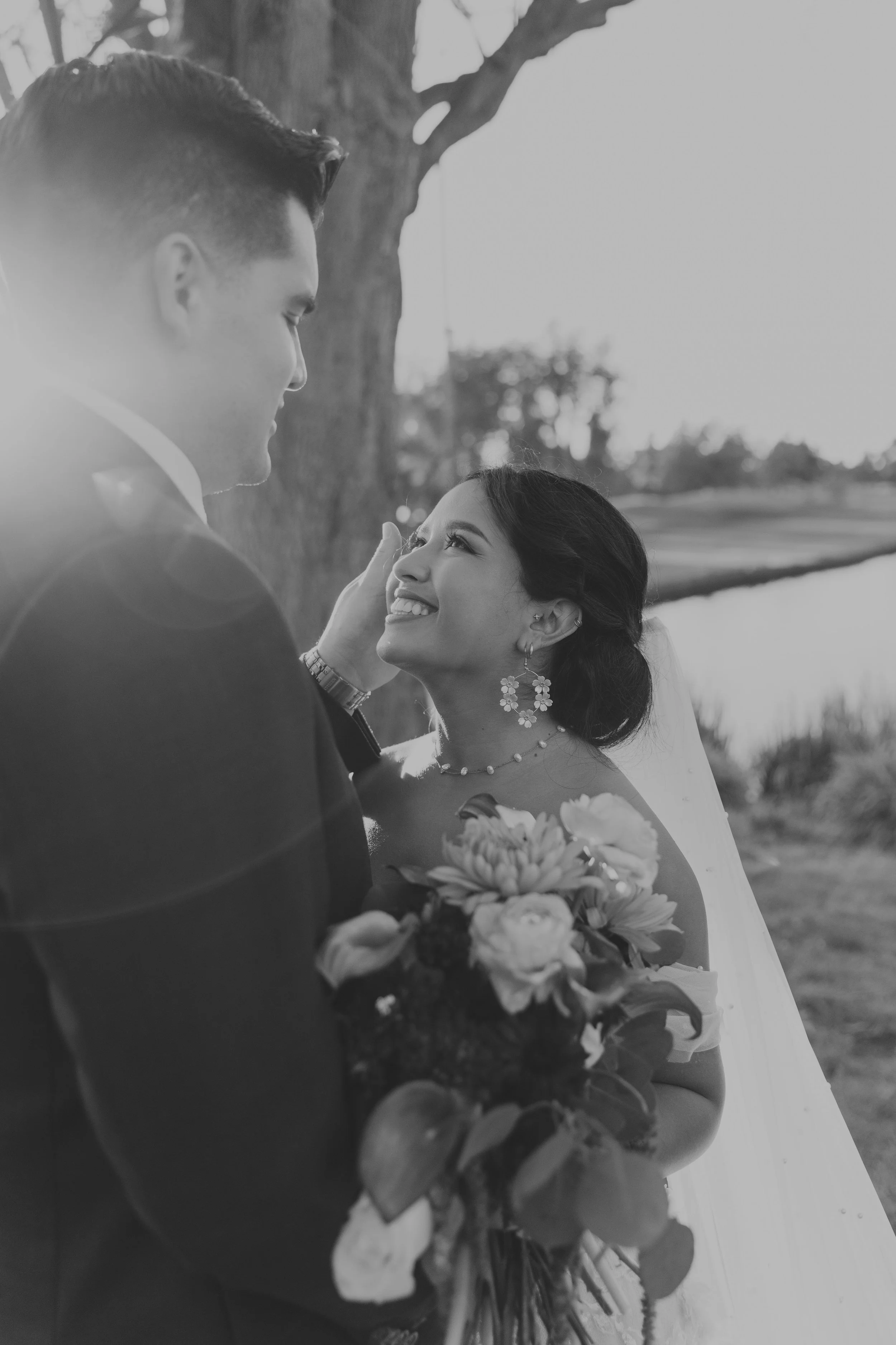 A black and white photo of a groom and bride sharing a joyful moment outdoors, with the groom touching the bride's face and holding a bouquet of flowers.
