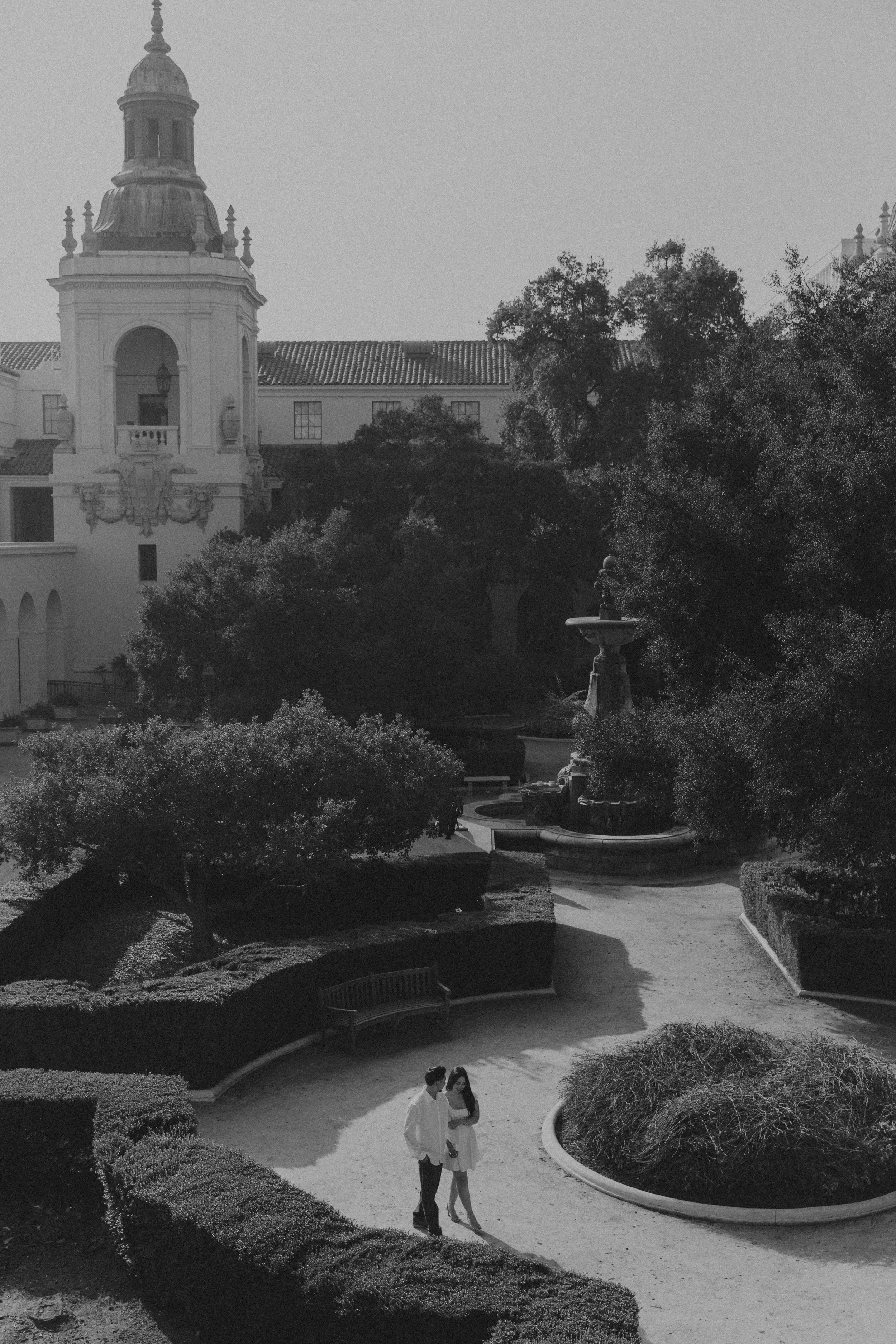 A black and white photo of a landscaped park with a fountain, trees, and a historic building with a clock tower in the background. Two people are walking along a curved pathway.