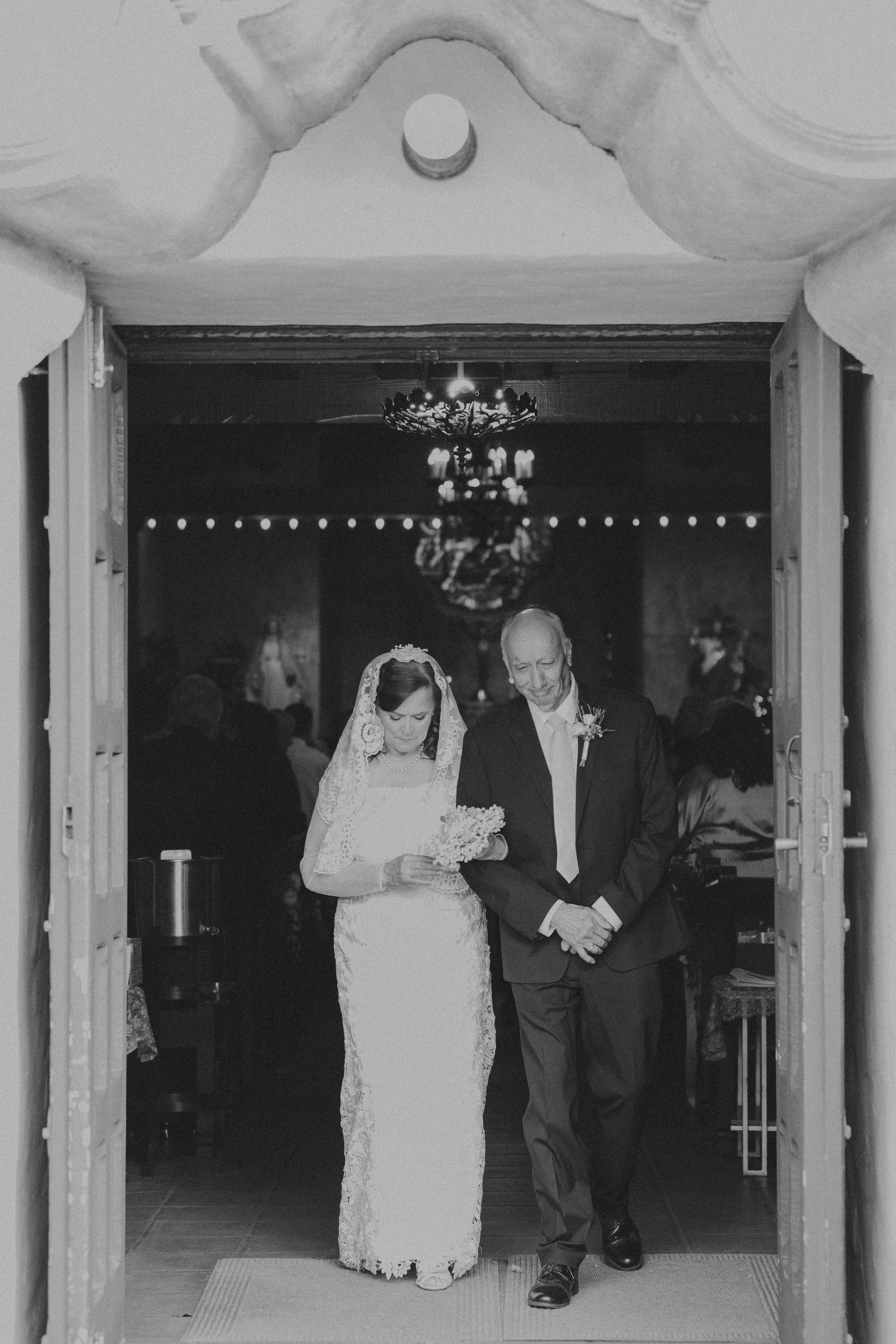 A bride and an older man walking down the aisle during a wedding ceremony, seen through the doorway of a church or chapel, with a chandelier hanging from the ceiling in the background.
