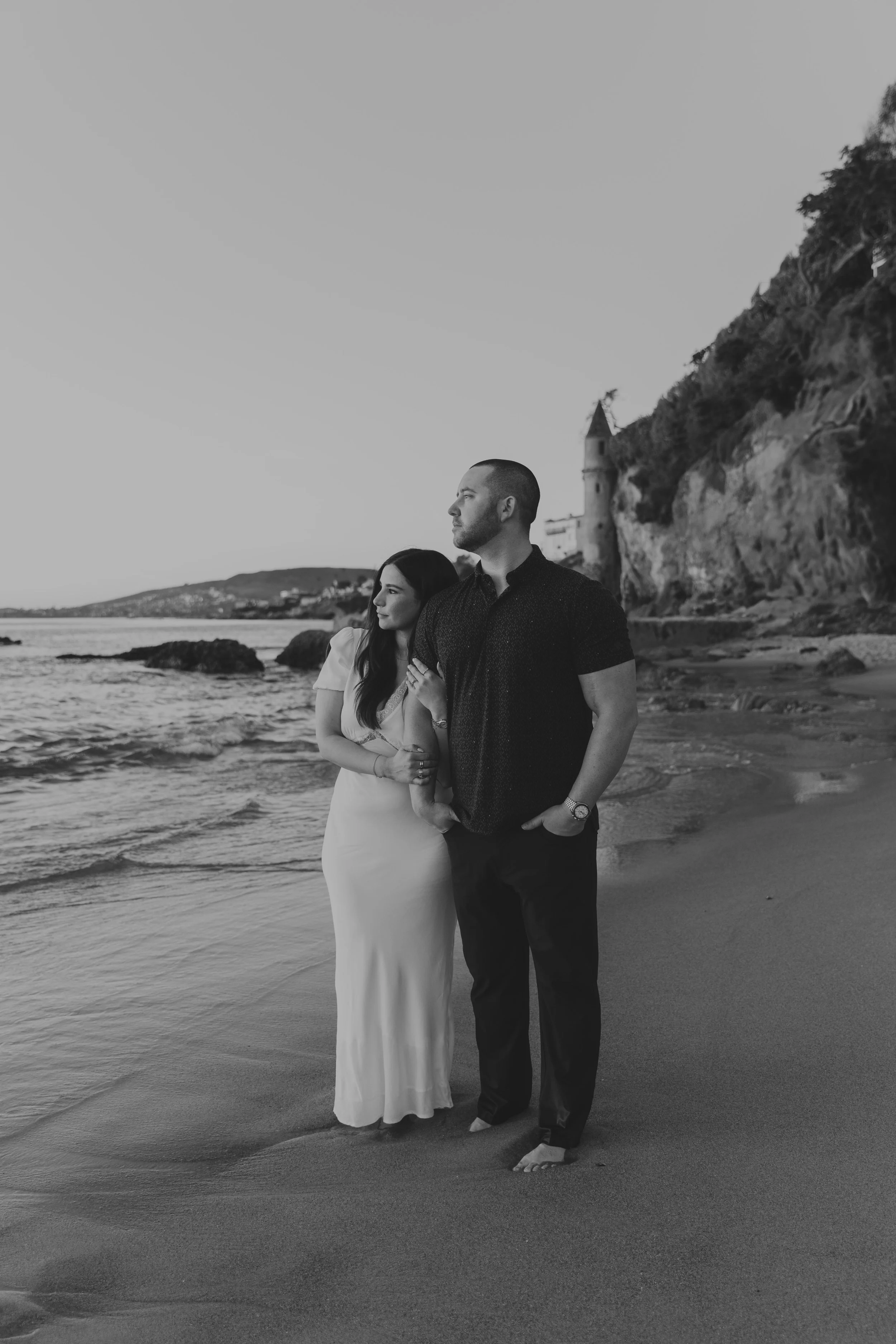 A black-and-white photo of a couple standing on the beach near the water, with rocky cliffs and a tower in the background. The woman is wearing a white dress and the man is dressed in dark clothing. They are holding hands and looking out at the ocean.