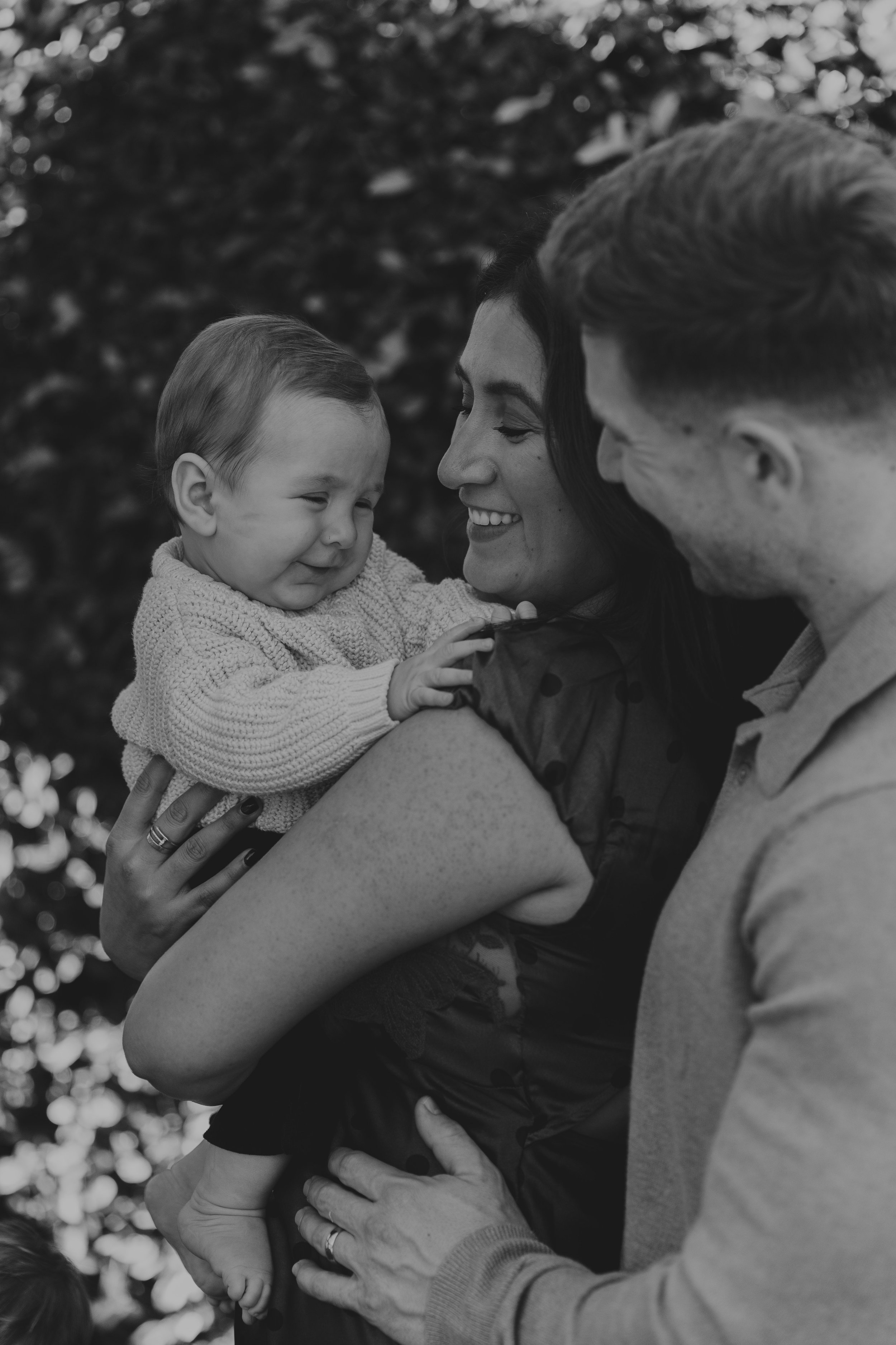 A black and white photo of a family celebrating a pregnancy. A woman, with a joyful expression, is holding a young child with Down syndrome. The child's hand is touching her face, and she is smiling. A man, likely the child's father, is close to the woman, smiling and looking at the child. The background is blurred with bokeh effects.