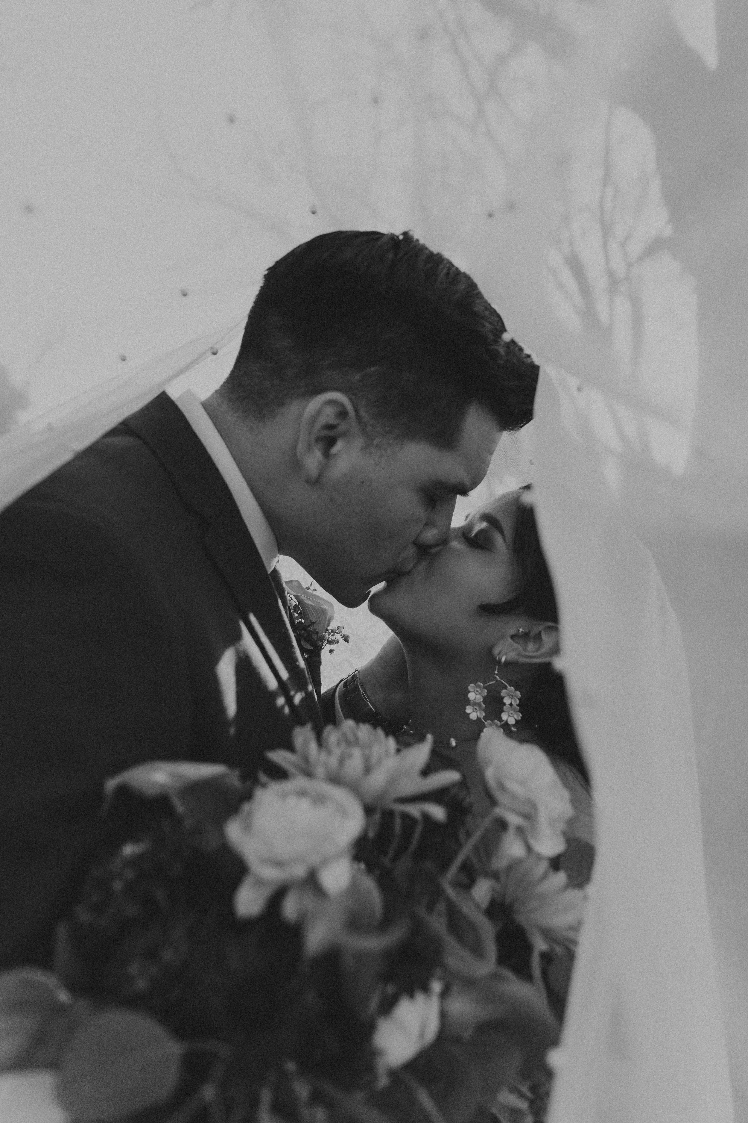 Black and white photo of a man and woman kissing at their wedding, with a bouquet of flowers in the foreground and trees in the background.