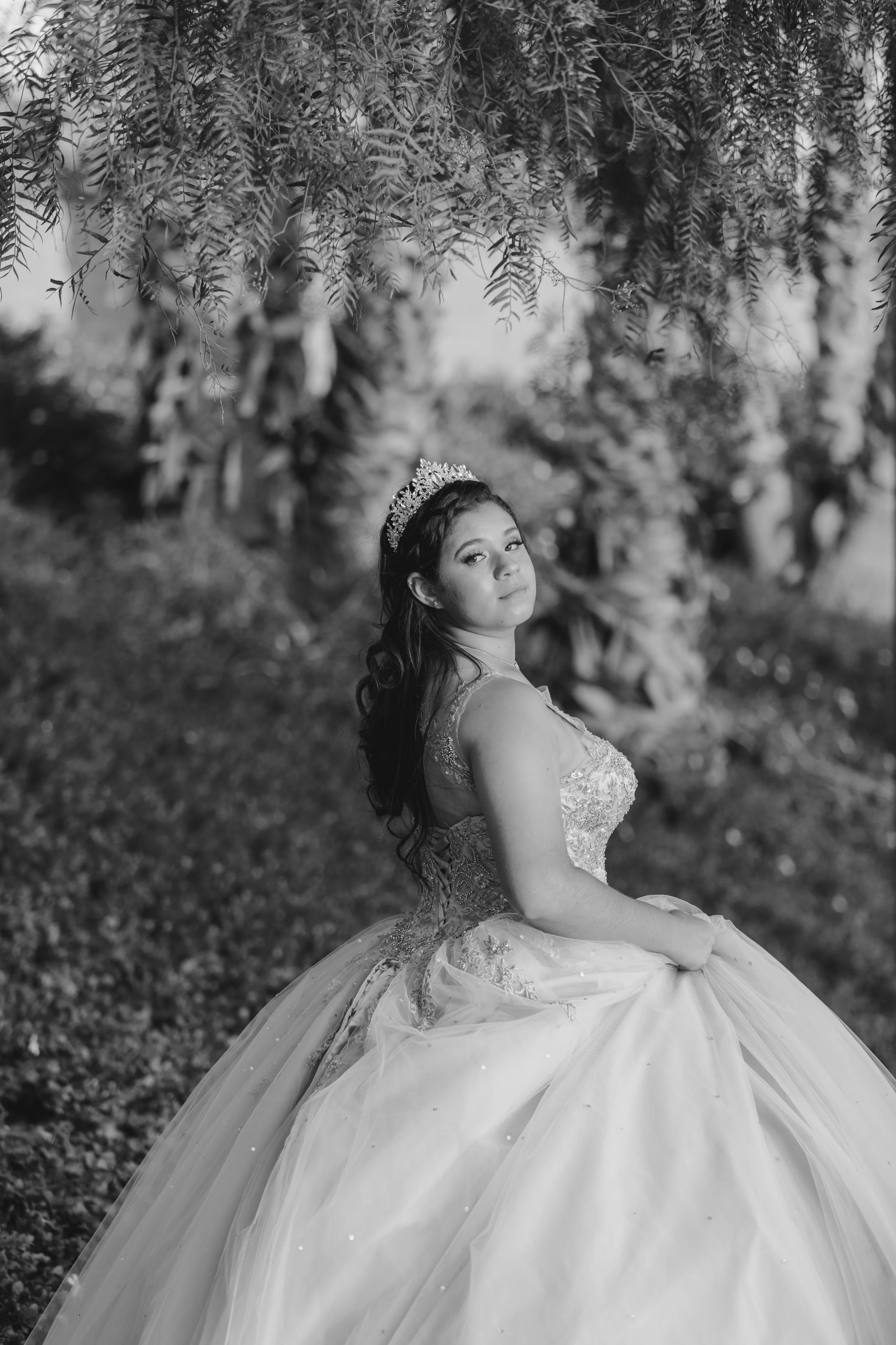 Black and white photo of a young woman in a wedding dress and tiara, sitting outdoors among trees with hanging branches.