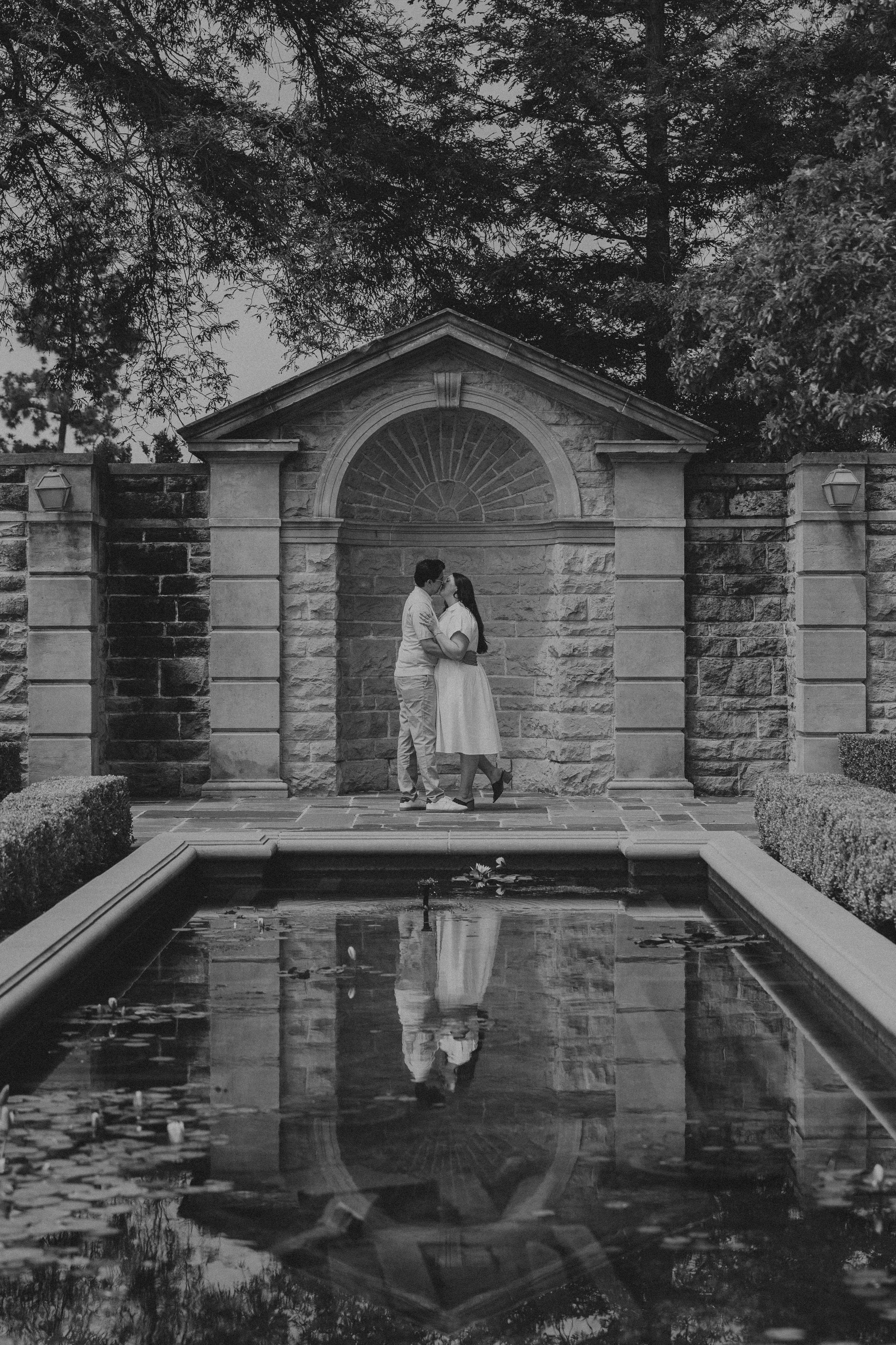 A black and white photo of a couple standing and kissing in front of a stone archway with trees overhead and a fountain reflecting the scene