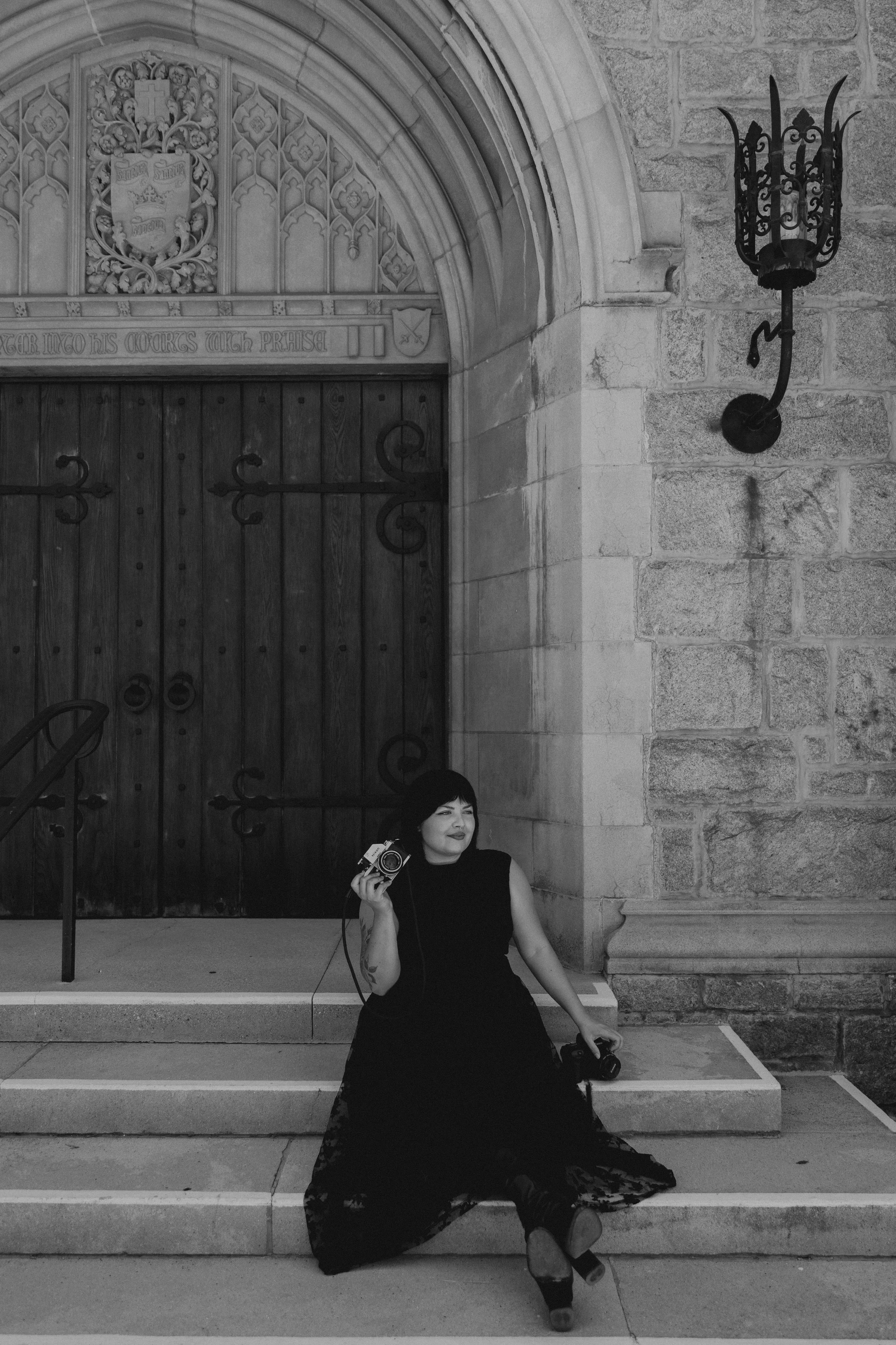A woman with dark hair, wearing a black dress, is sitting on the steps outside a stone building with an arched doorway. She is holding a vintage camera and smiling, with a second camera on the ground beside her. The building features intricate stone carvings and a wrought-iron light fixture on the wall.