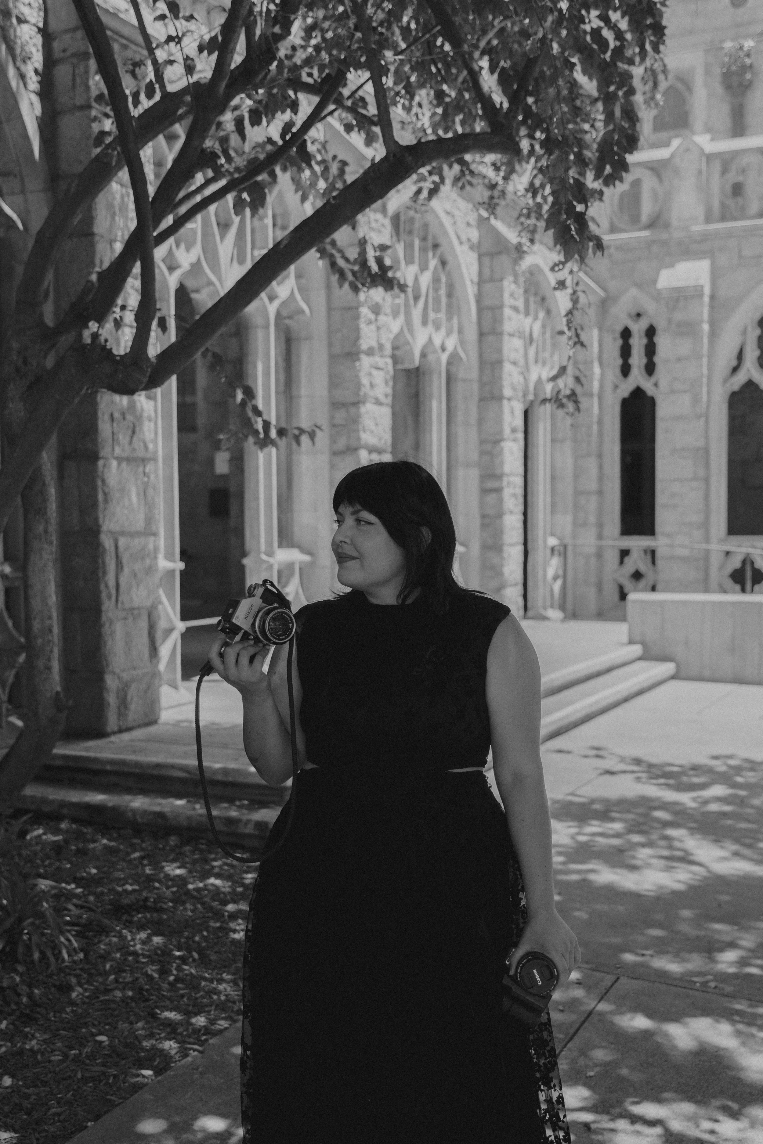 A woman in a black dress holding a camera and a drink stands outdoors at night in front of an old stone church, with trees and benches around her.
