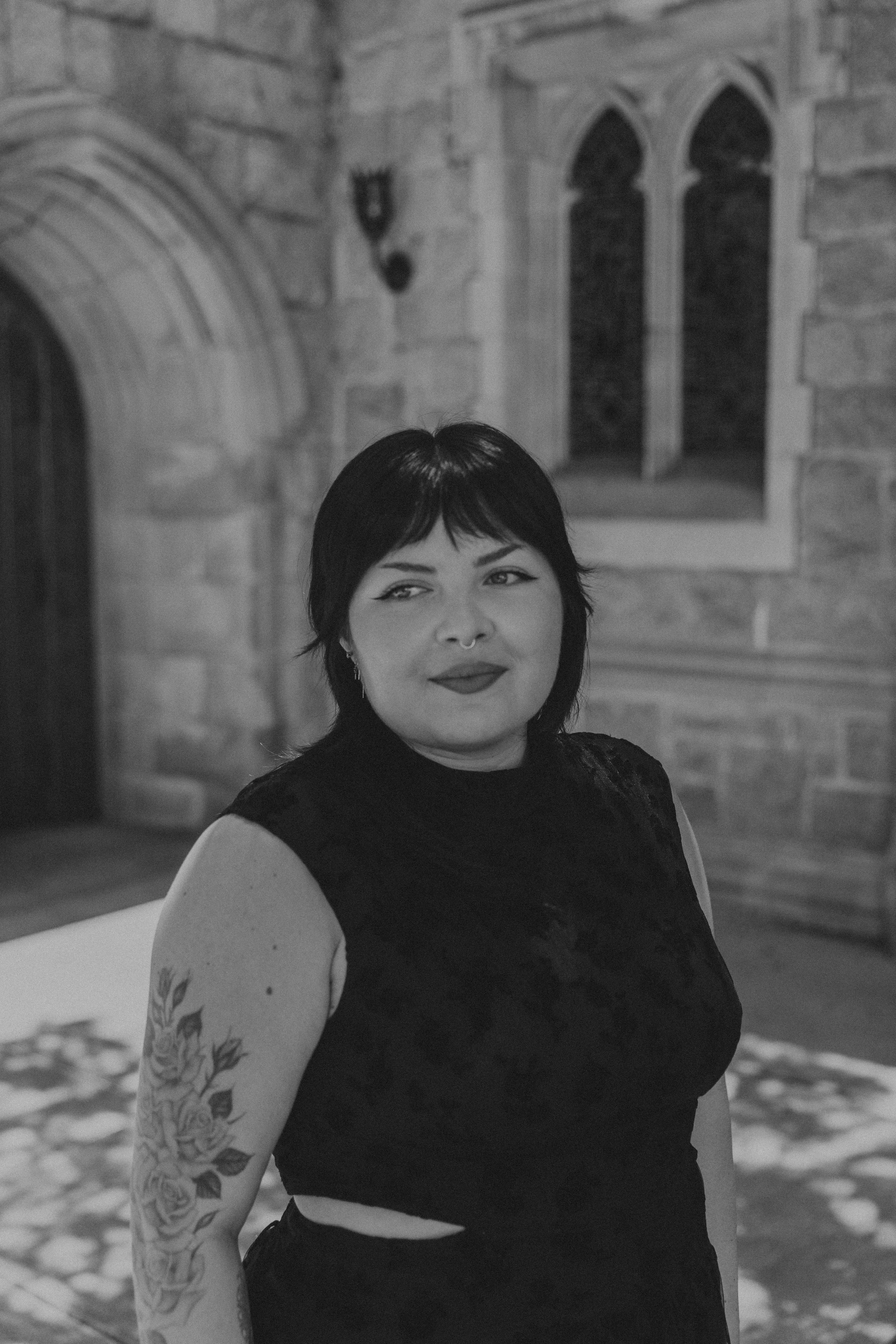 A woman with short dark hair and tattoos on her arm, wearing a black sleeveless top, standing outdoors near a stone building with gothic windows and a curved entrance.