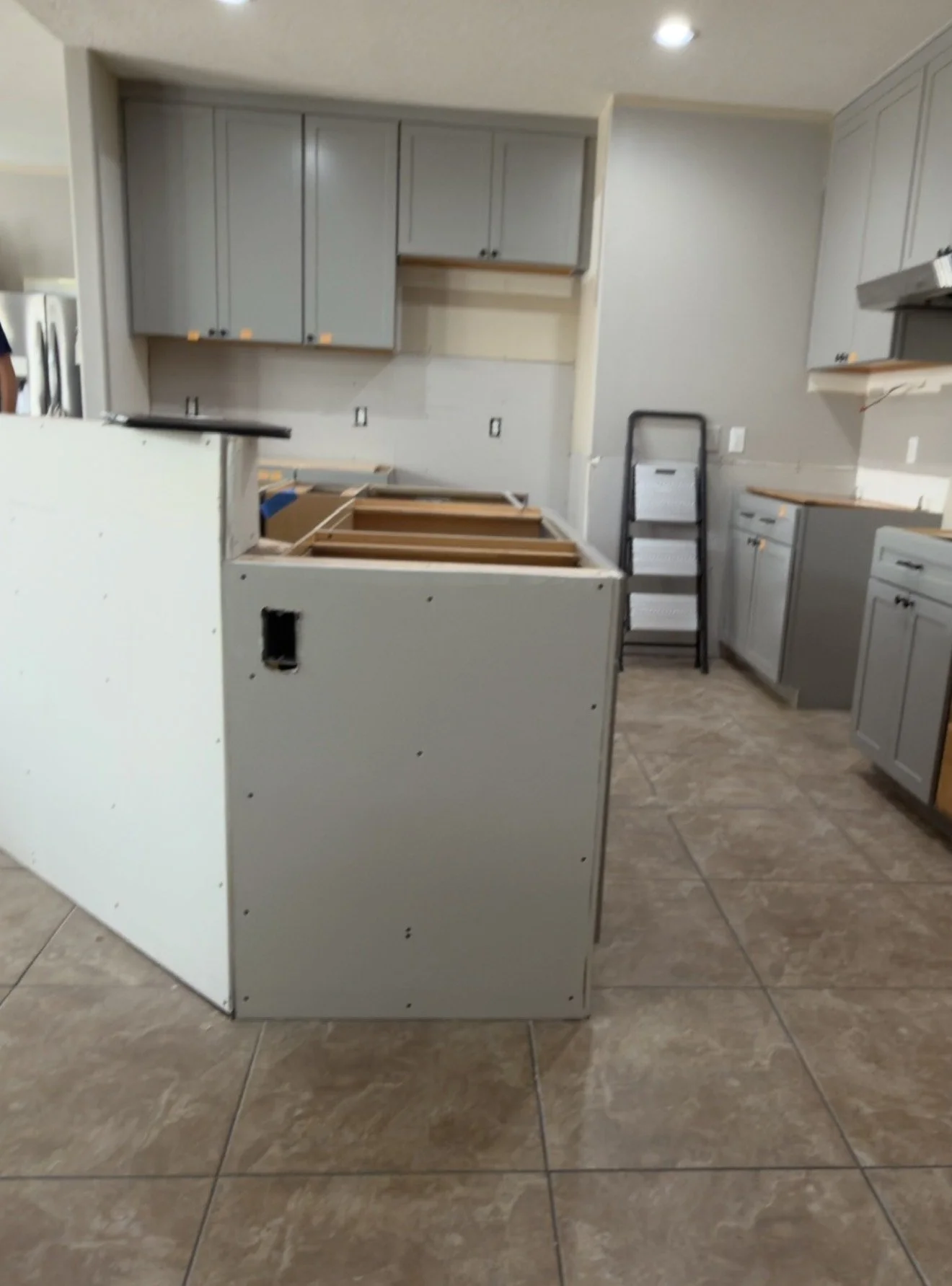 Kitchen under renovation with partially installed cabinetry and an unfinished island, featuring upper and lower cabinets in a light color, a folded step ladder, and tiled floor.