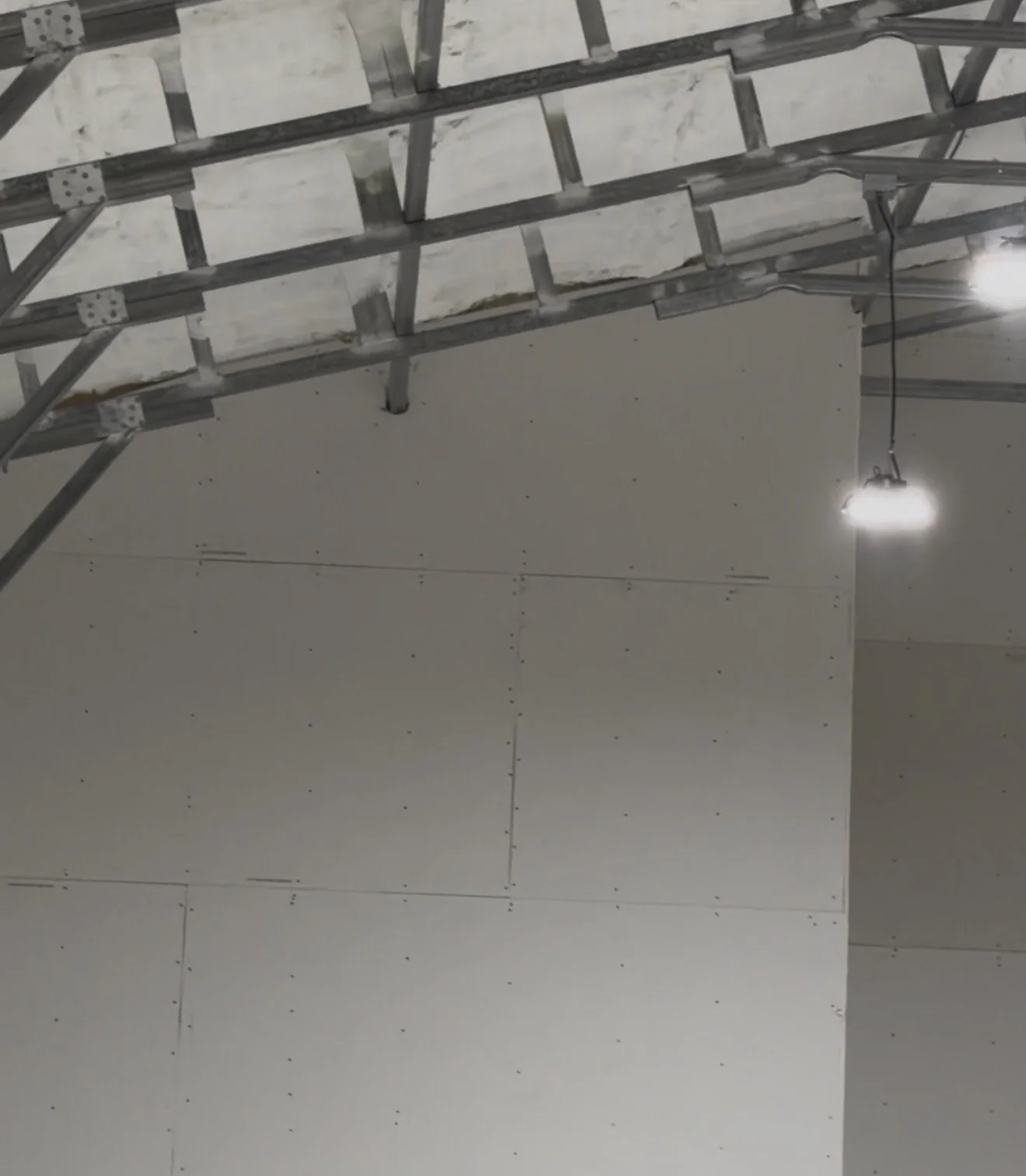 Interior of a room under construction with drywall panels on the ceiling and a metal scaffolding structure. There is a bright overhead light hanging from the ceiling.