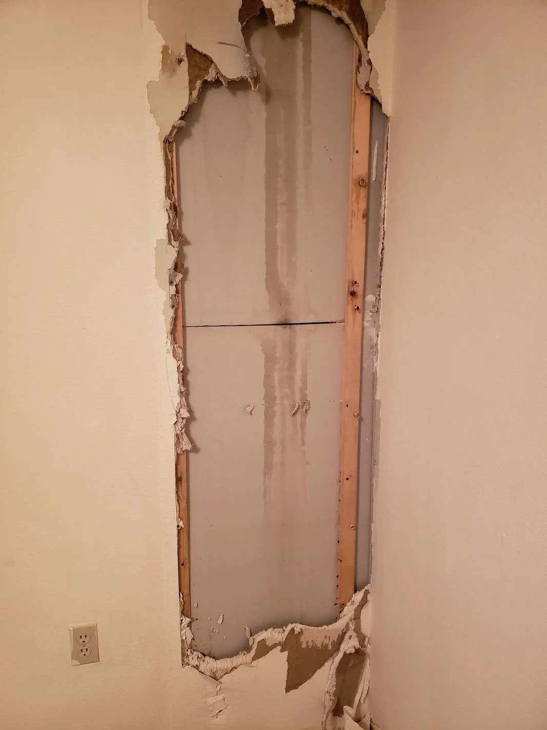 Open wall with drywall removed, revealing wooden framing and electrical outlet, likely for plumbing or electrical work.
