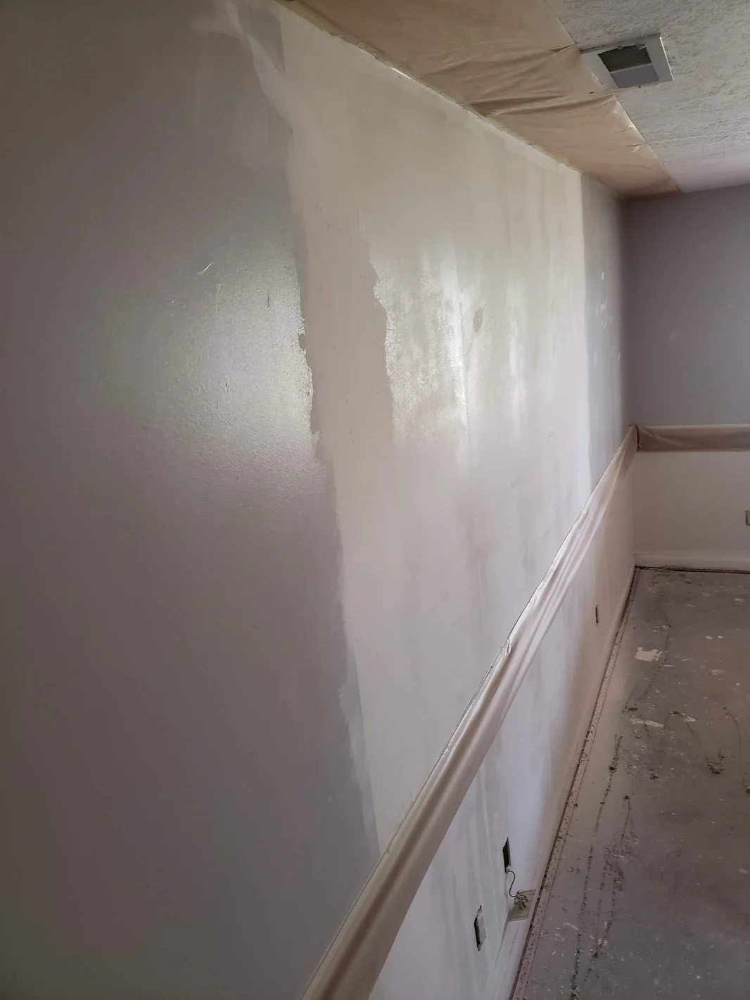 Interior of a room with an unfinished wall, partially painted white and beige tape along the ceiling, with visible electrical outlets and a vent.