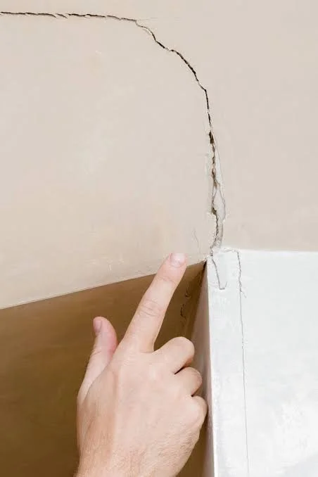 Person pointing at a crack in the wall near the corner where the wall meets the baseboard.