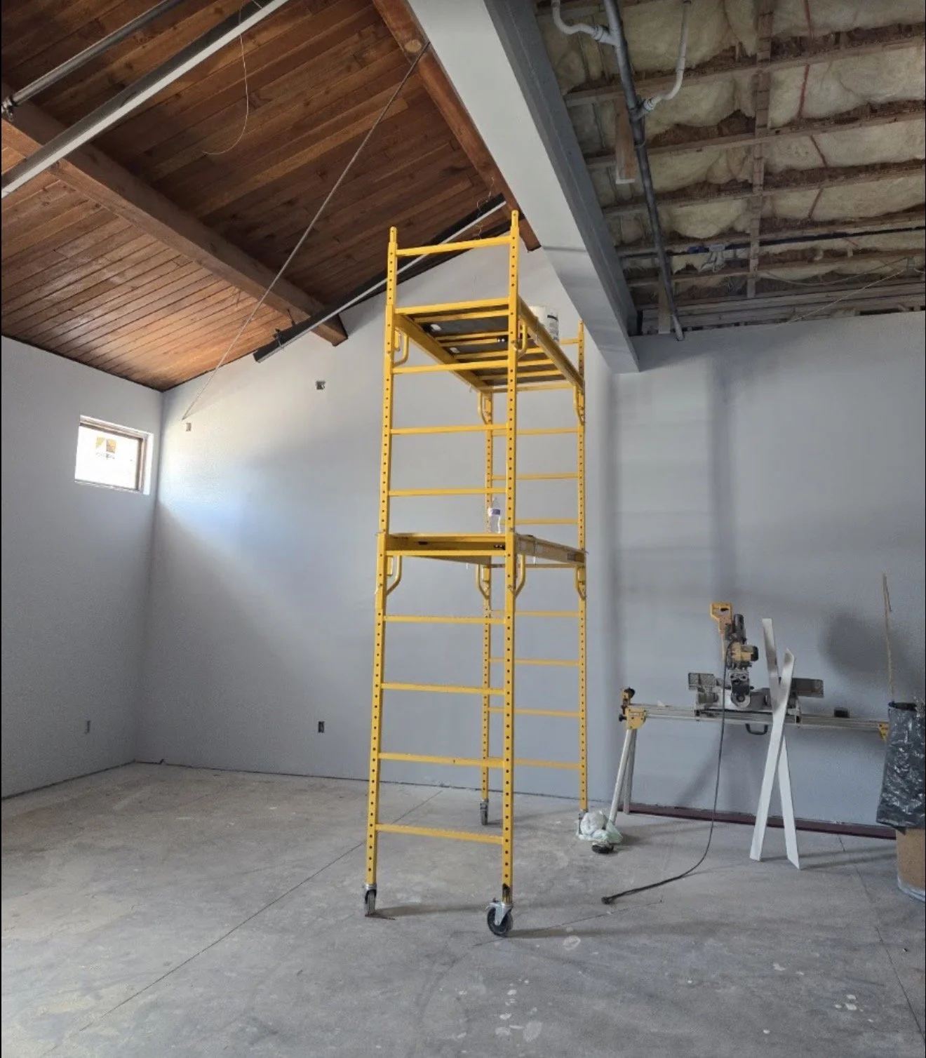 Construction site with a yellow rolling scaffold in the center, a partially finished ceiling with exposed insulation and pipes, a small window on the left wall, and construction tools on the right side.