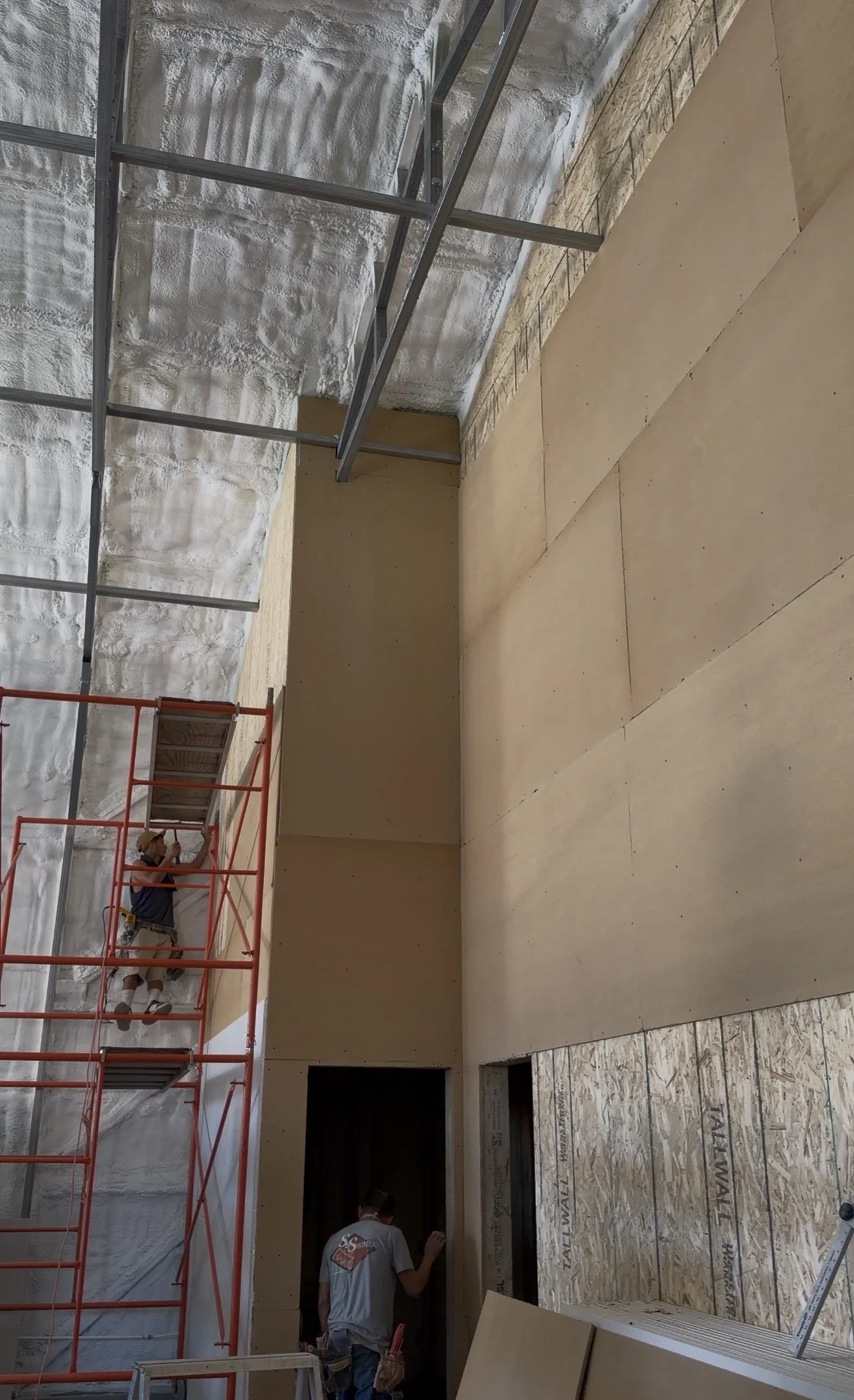 Construction workers installing drywall and insulation on an interior wall inside a building under construction.